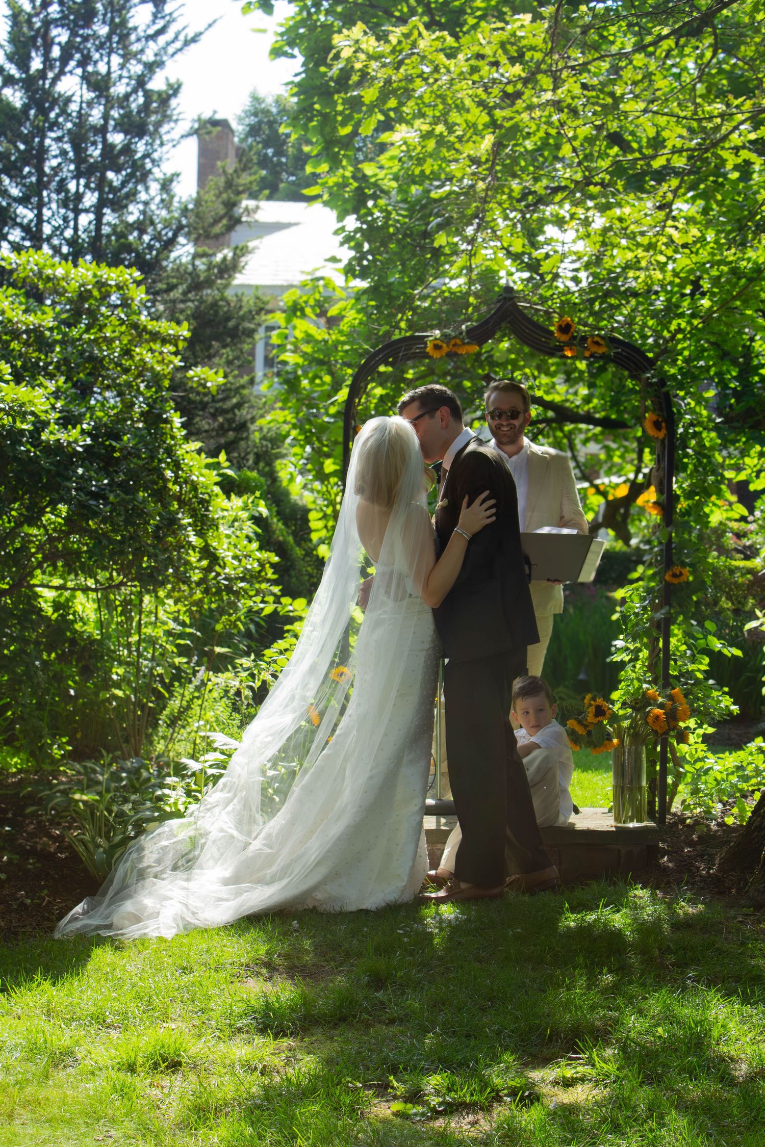 Bride and groom kissing under a rustic floral arch during an outdoor garden wedding ceremony on a sunny day.