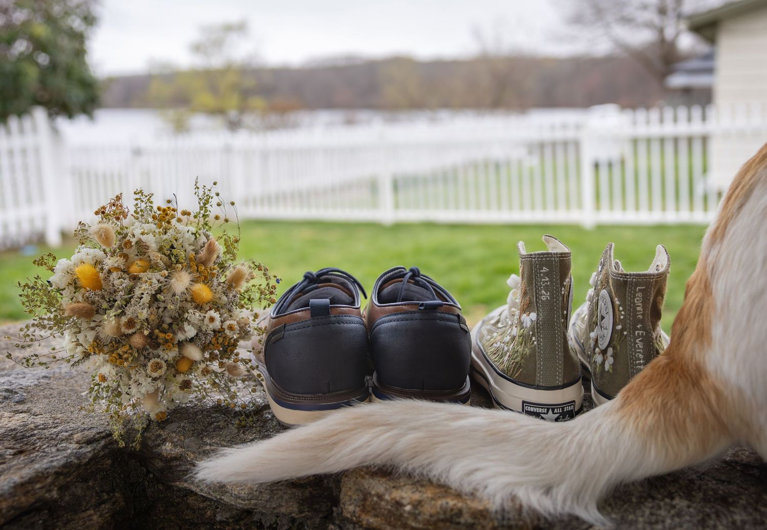 Wedding bouquet of dried yellow flowers with bridal shoes and boots on stone ledge near white fence.