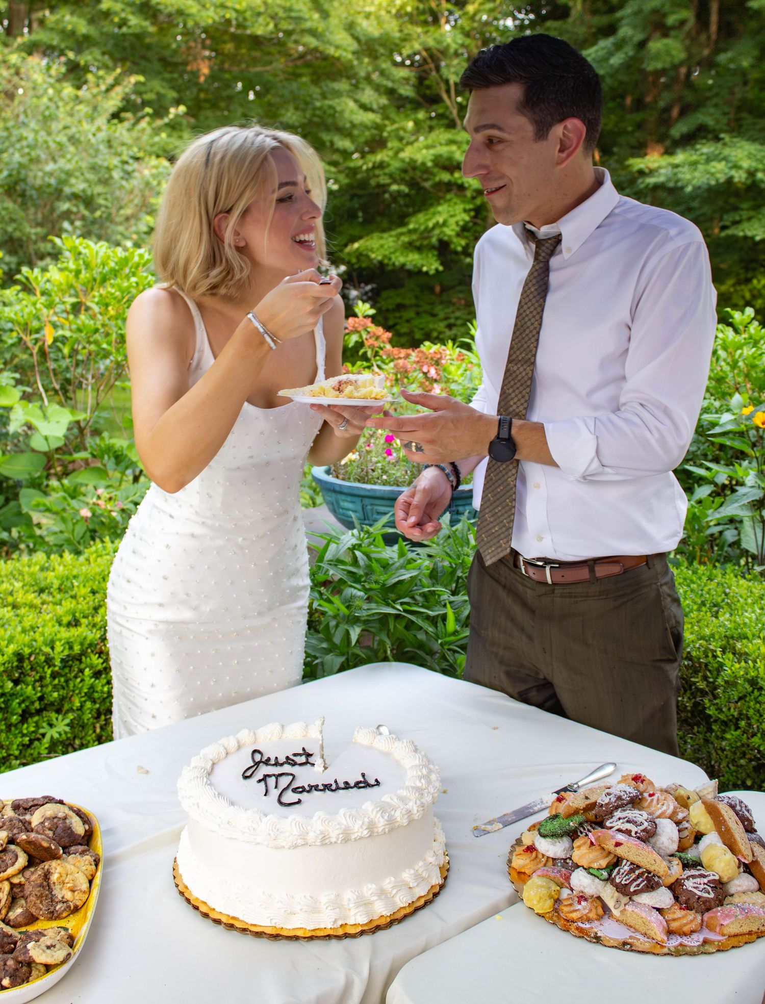 Bride and groom sharing cake at outdoor wedding reception with decorated dessert table.