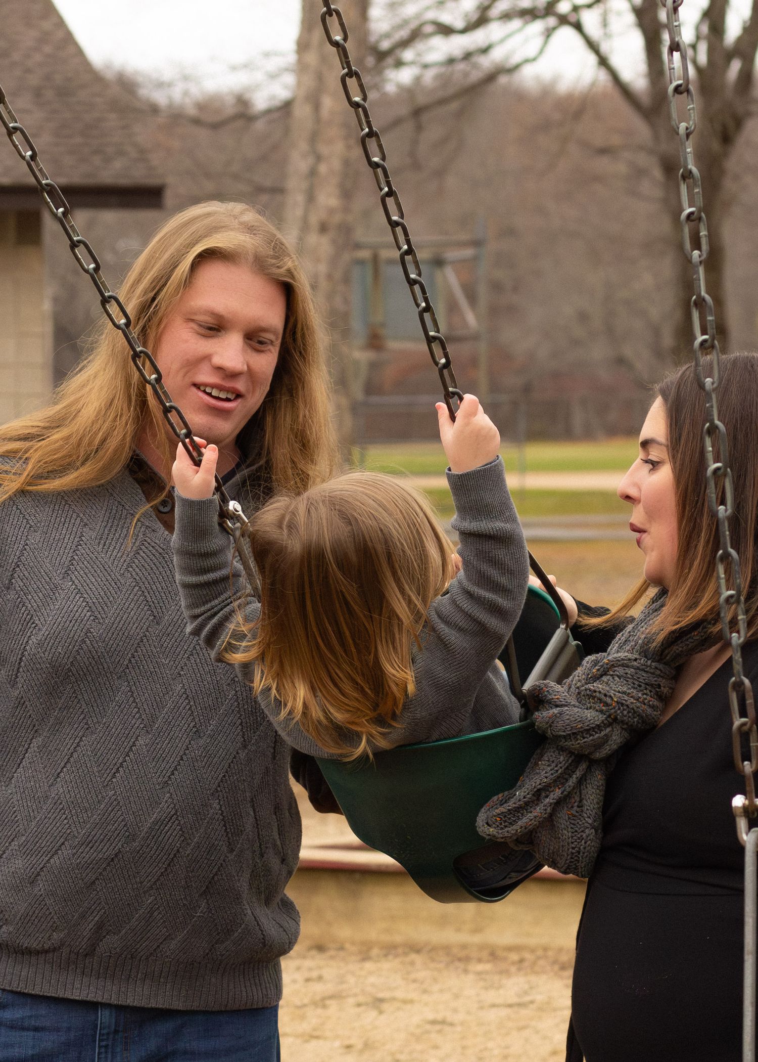 A young family with long-haired parents playing with their toddler on a green swing at a park playground.
