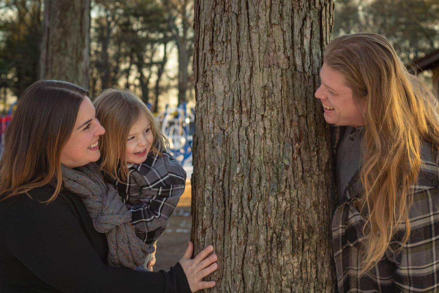 Happy family of three peeking around a tree trunk in a sunny park, sharing a joyful moment together.