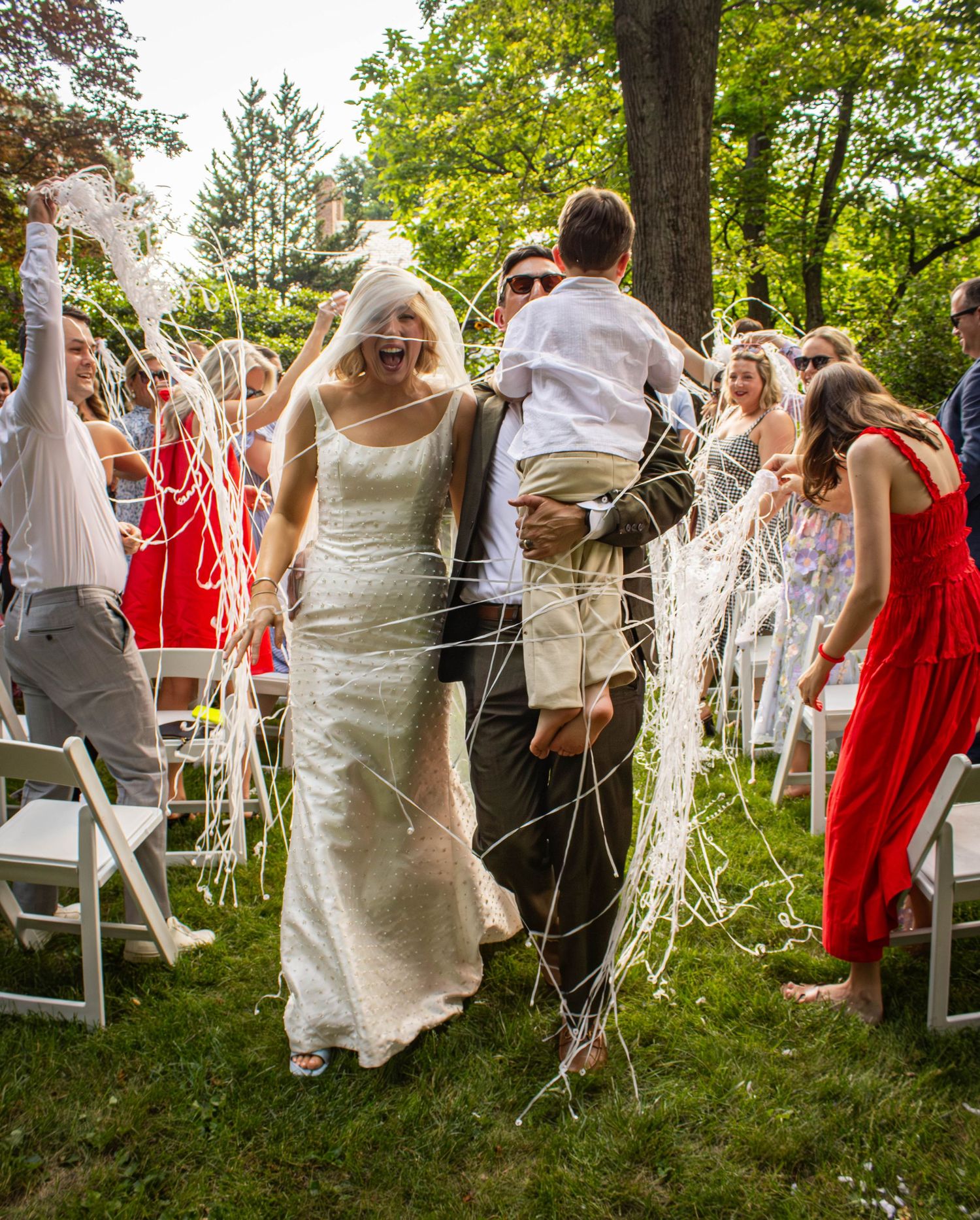 Joyful bride and groom walk down outdoor aisle as guests celebrate with streamers at a sunny garden wedding.