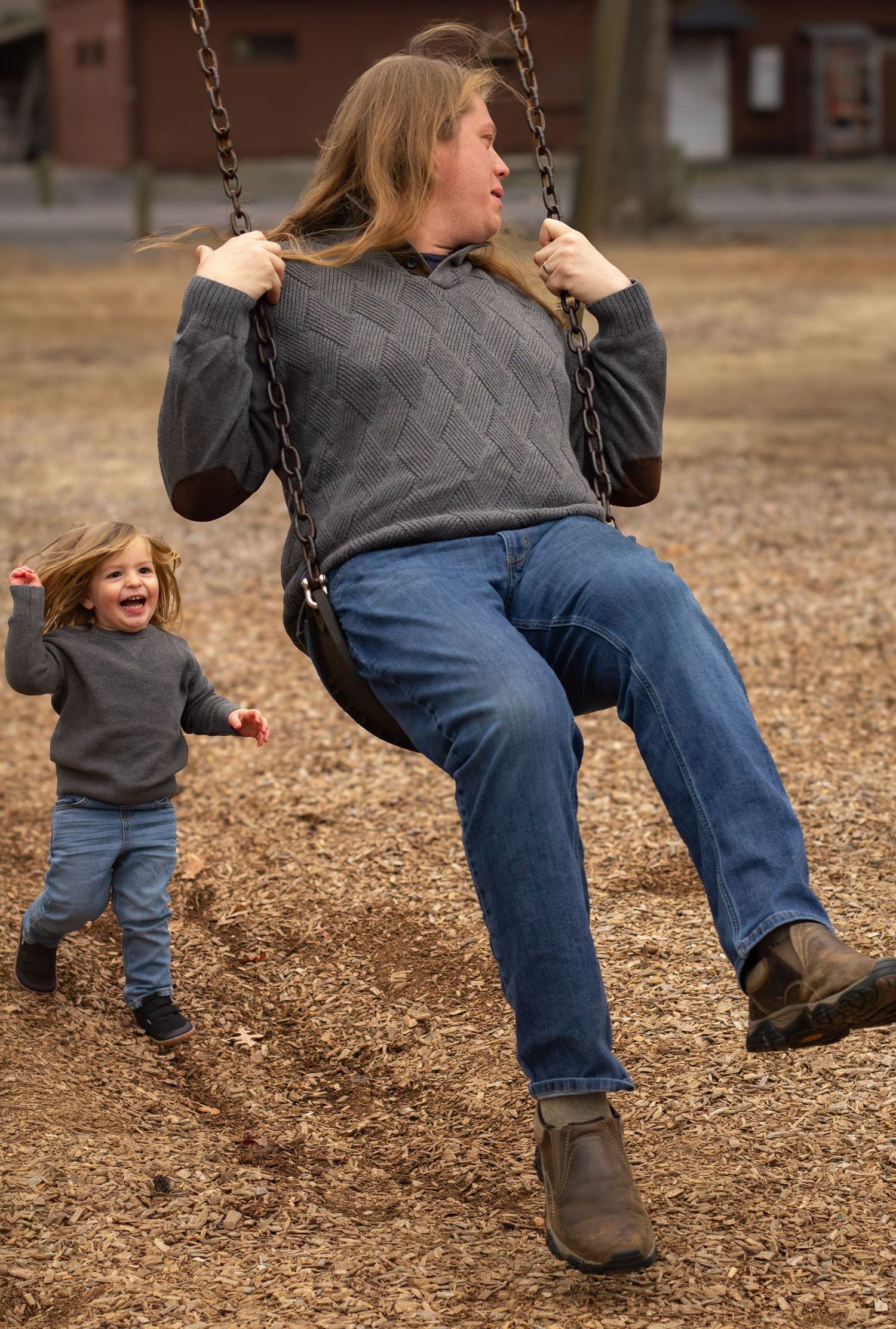 Smiling mother rides playground swing while laughing toddler runs behind her at outdoor park.