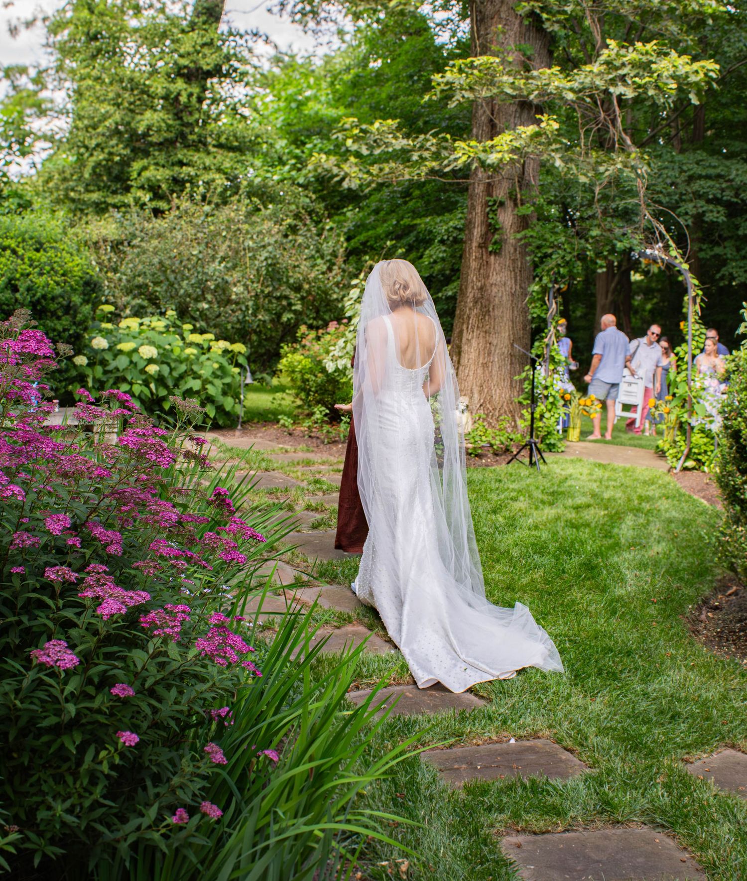Bride in white gown and veil walking along garden stone path surrounded by pink flowers and lush greenery.