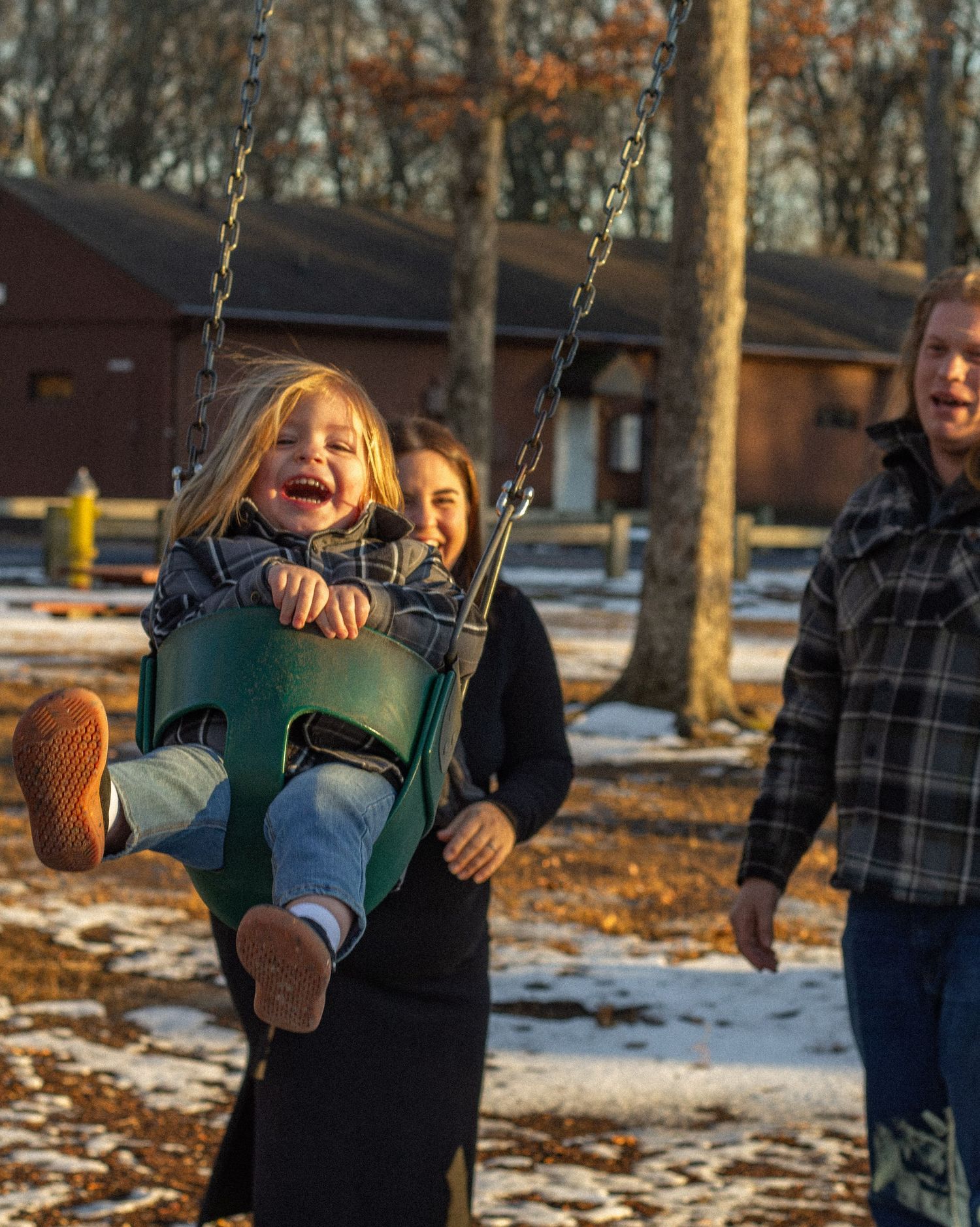 Joyful child laughing on a green swing while parents push him at a snowy winter playground.