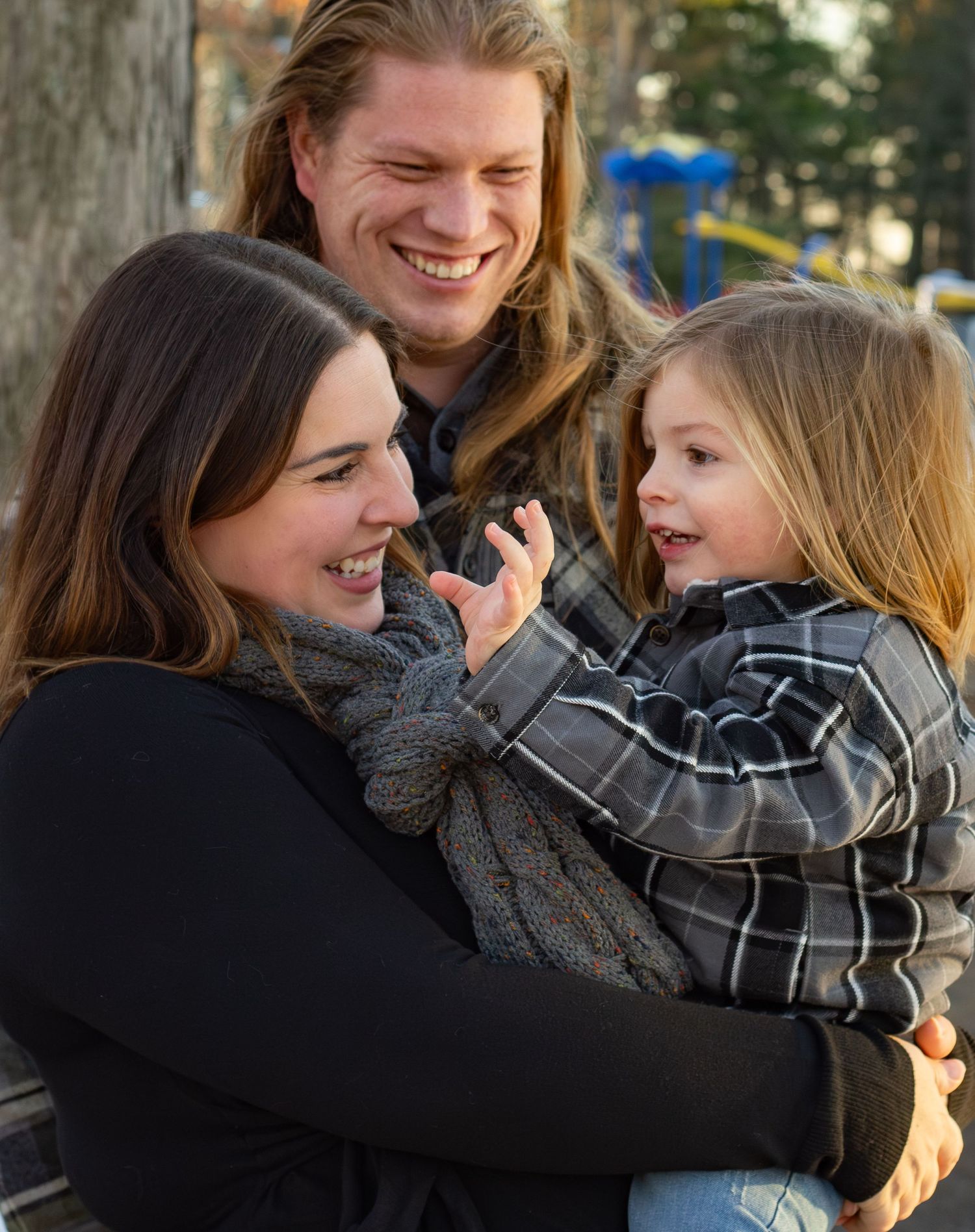 Laughing family portrait with parents and young child in plaid shirt sharing a joyful moment outdoors.
