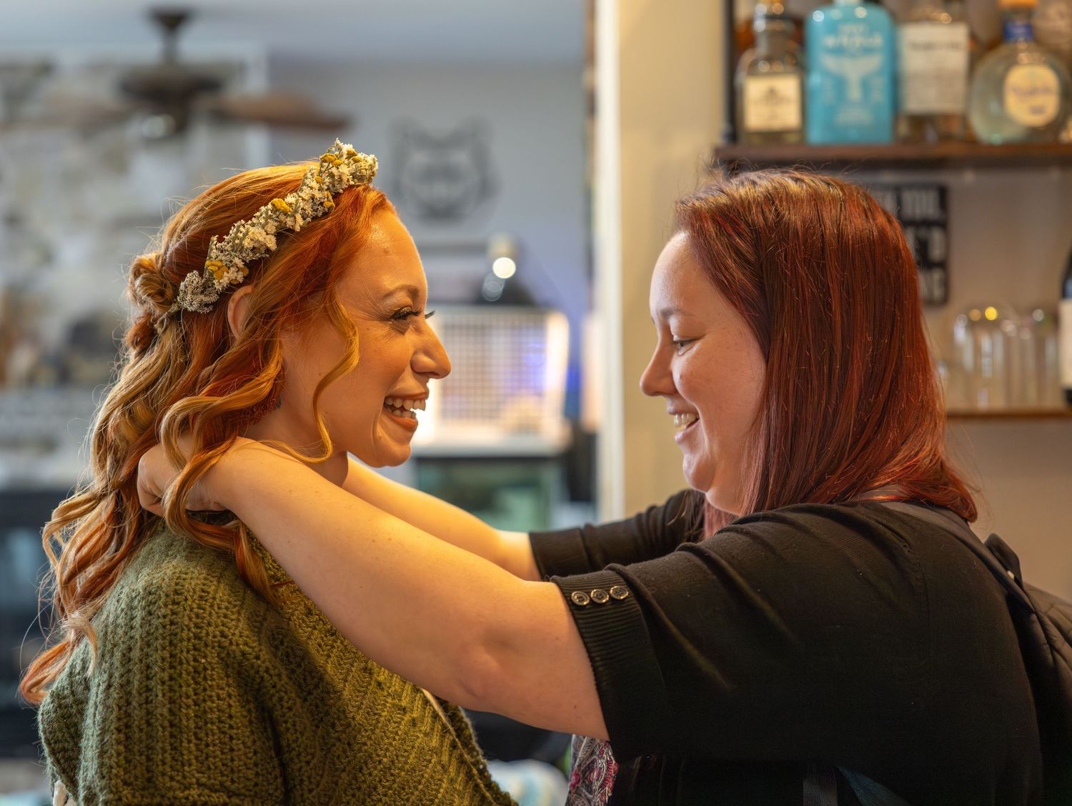 Two smiling women sharing a joyful moment as floral crown is placed on brides head.