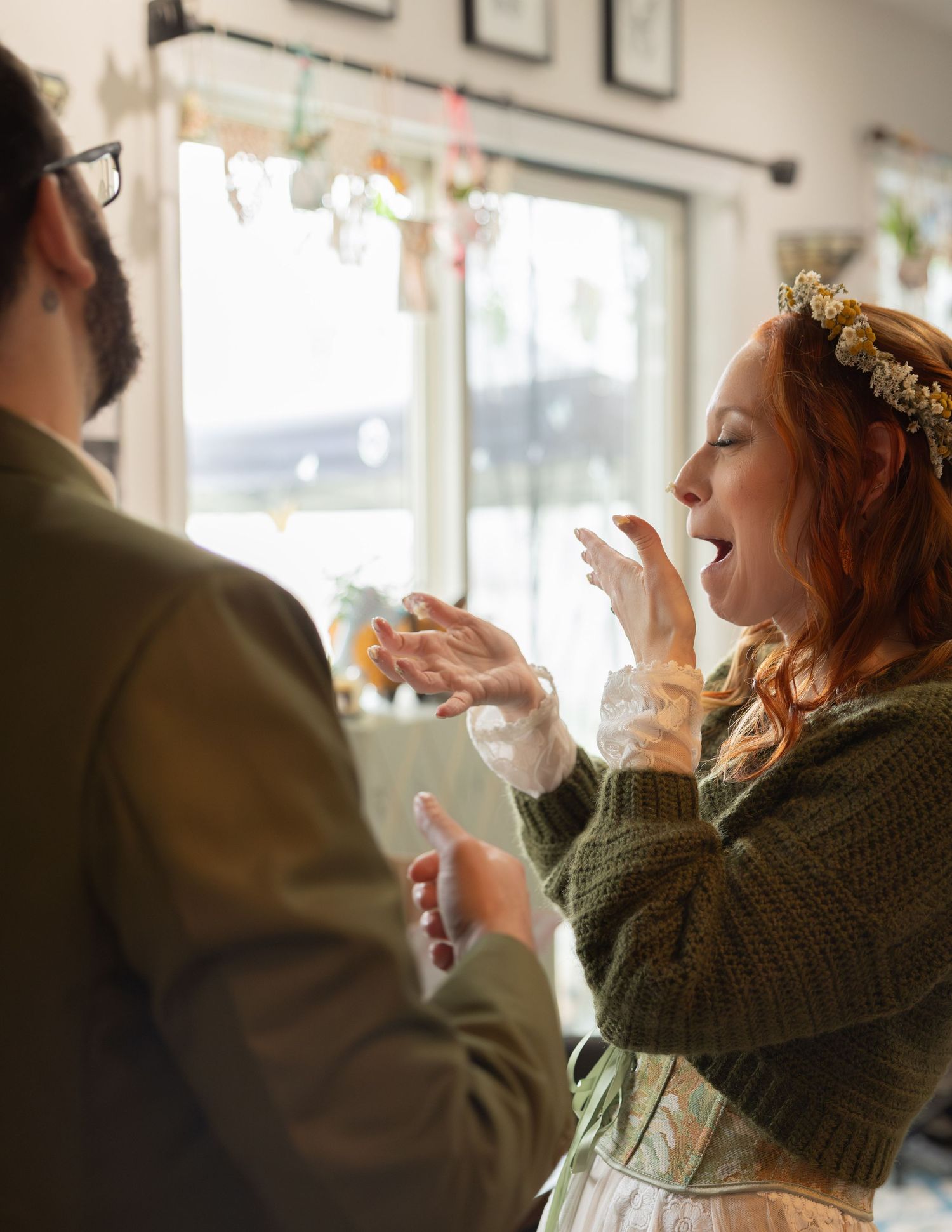 Excited bride in green sweater and floral crown reacts joyfully during wedding celebration indoors.