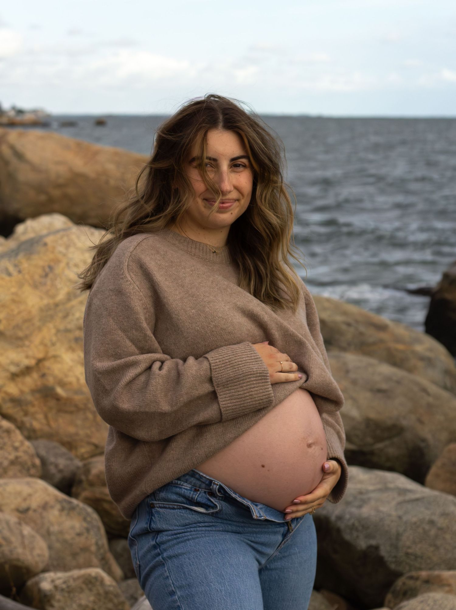 Beautiful pregnant woman in brown sweater cradling baby bump while posing on rocky ocean shoreline.
