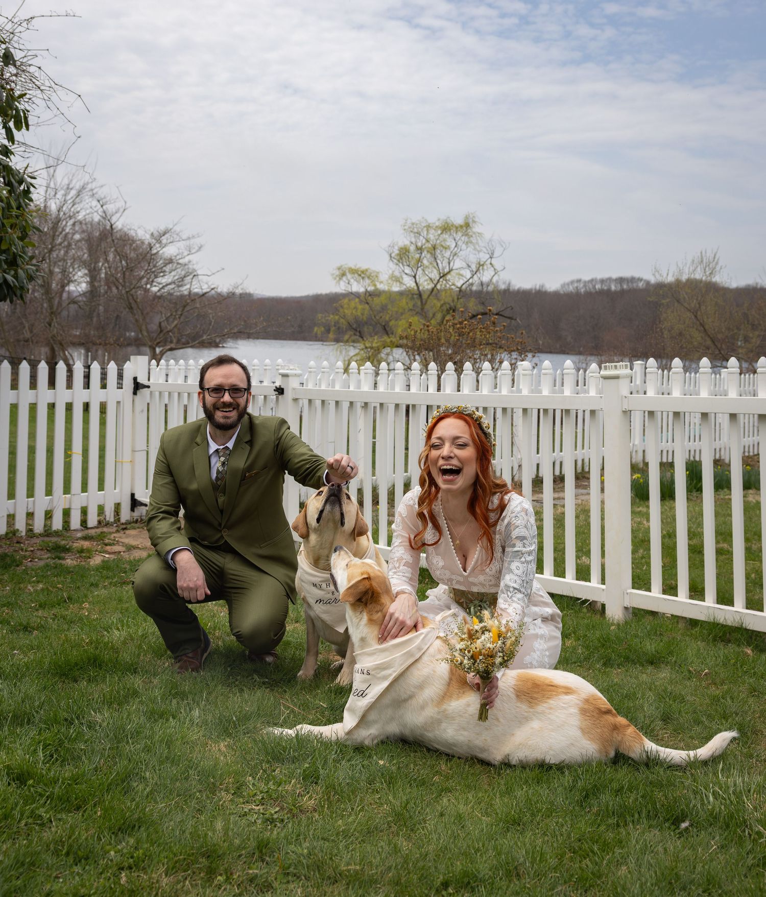 Laughing bride and groom in green suit pose with golden retriever dog by white picket fence outdoors.