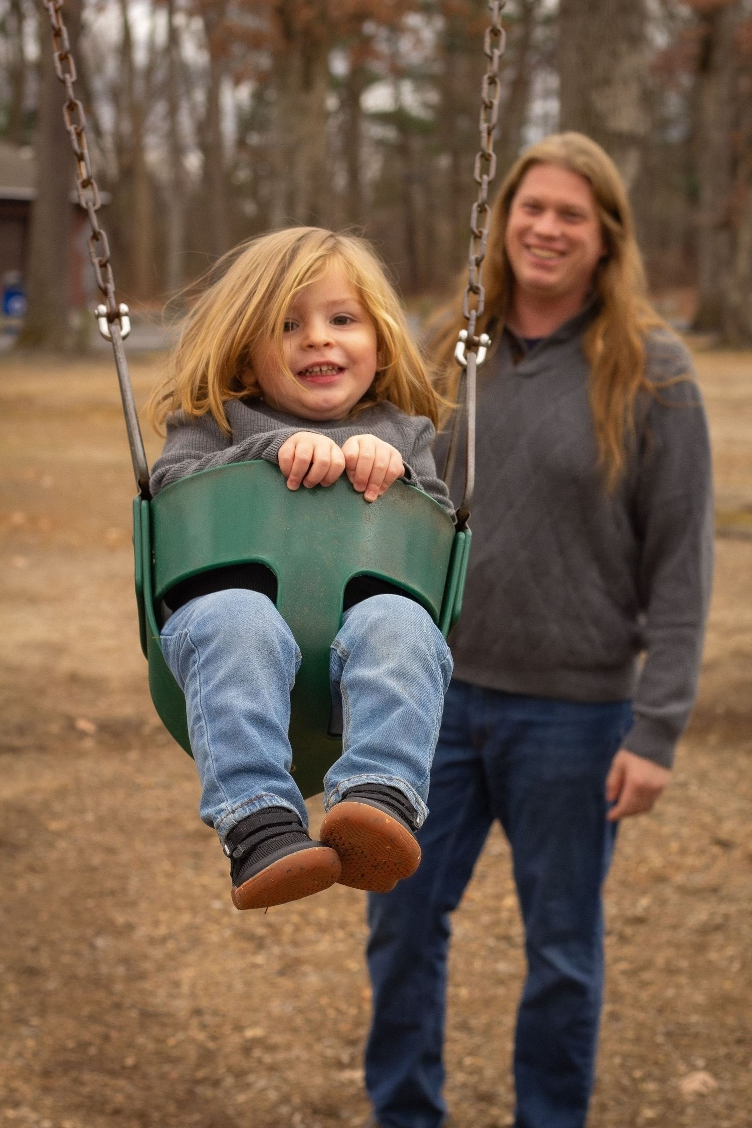Happy young child swings in green bucket swing while smiling long-haired parent pushes from behind.