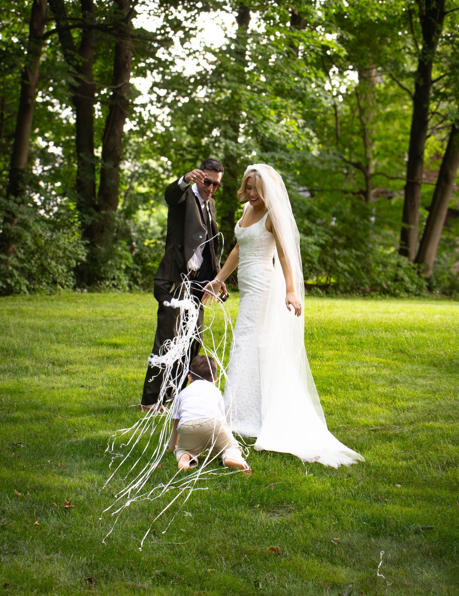Bride and groom stand on green lawn while child plays with white ribbon streamers during outdoor wedding celebration.