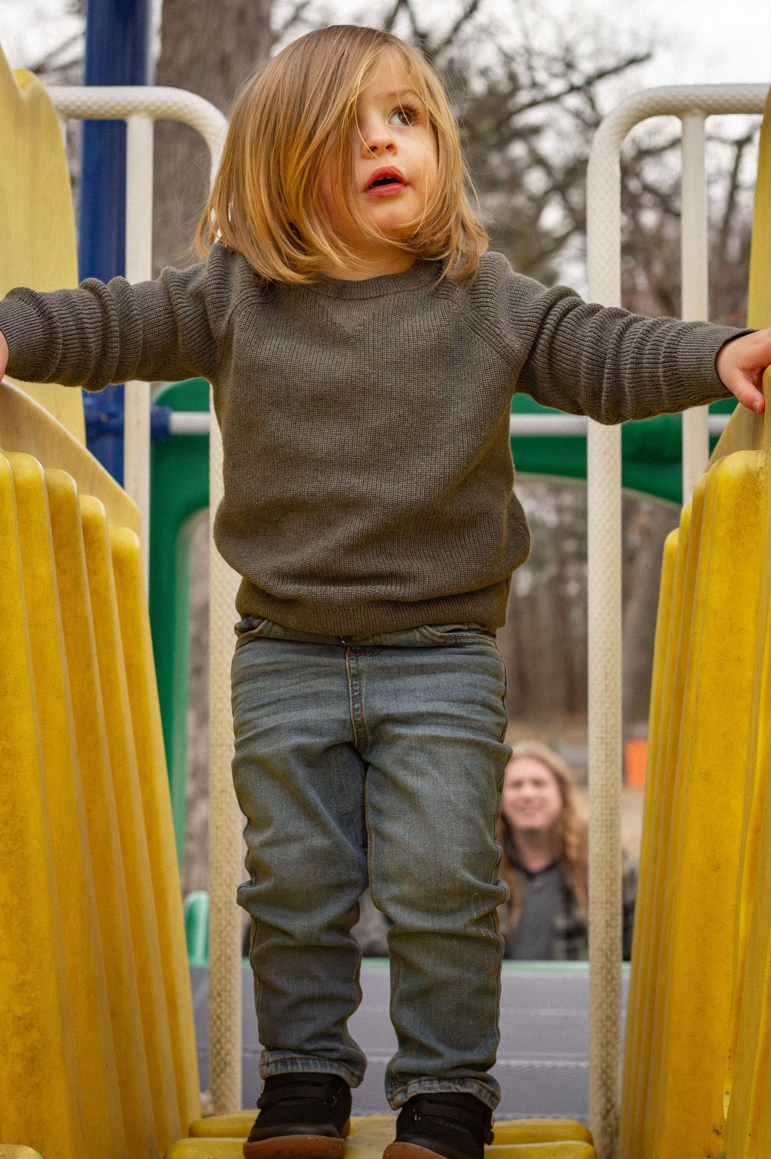 Adorable toddler with blonde hair standing on yellow playground equipment wearing jeans and brown boots.