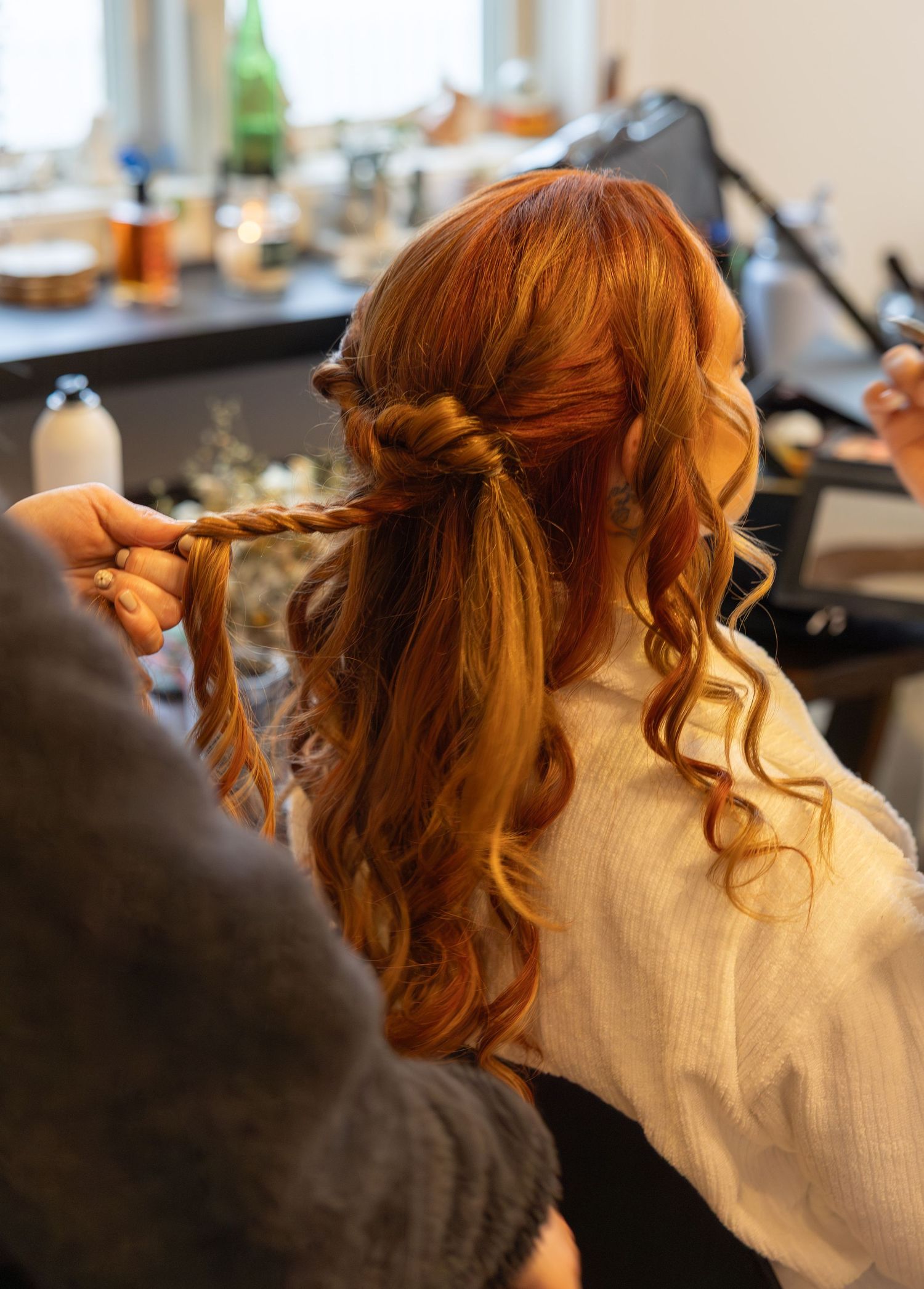 Stylist braiding long red wavy hair of bride during wedding preparation in salon setting.