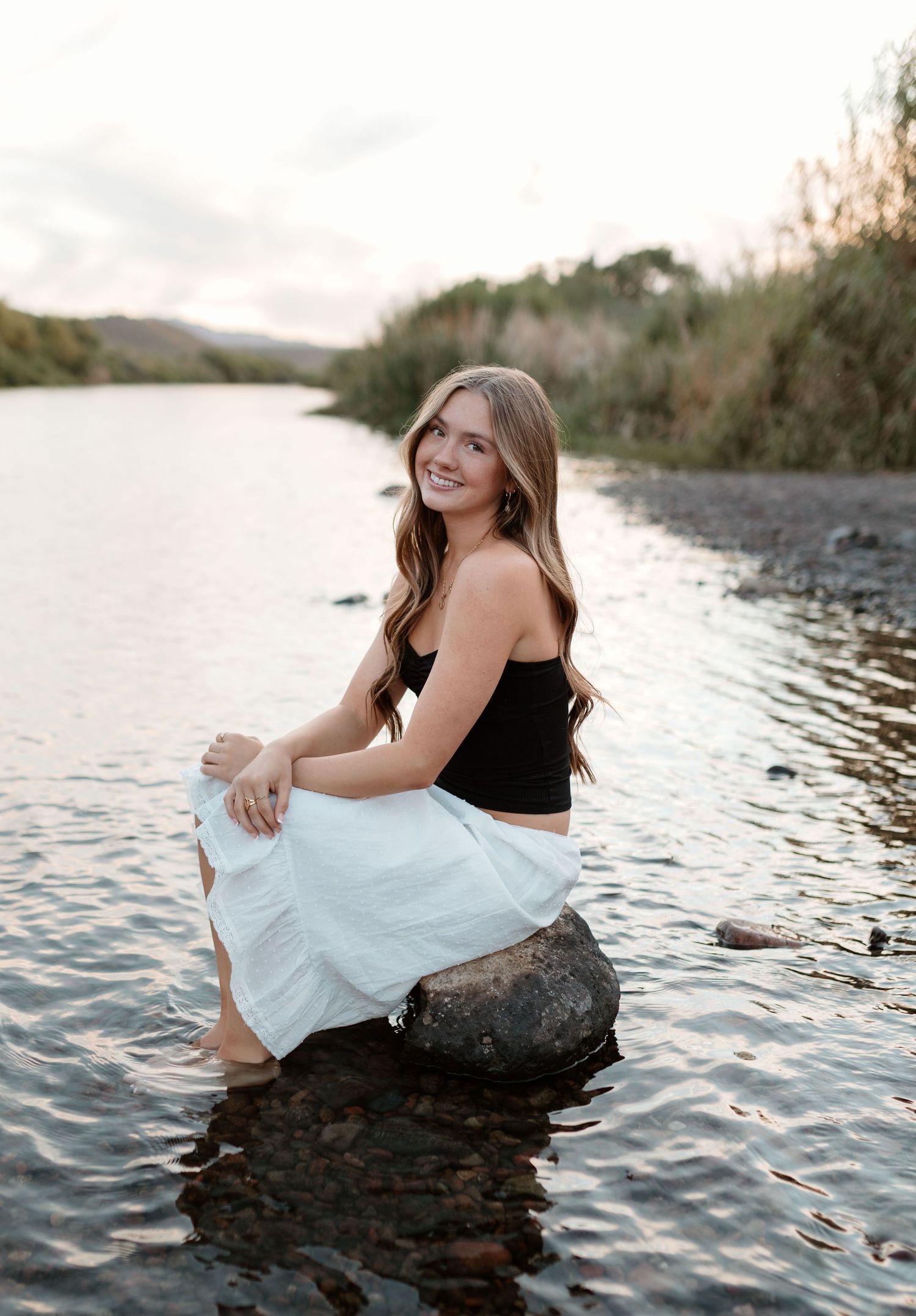 Young woman in black crop top and white skirt sitting on a rock in a shallow river, smiling at sunset.