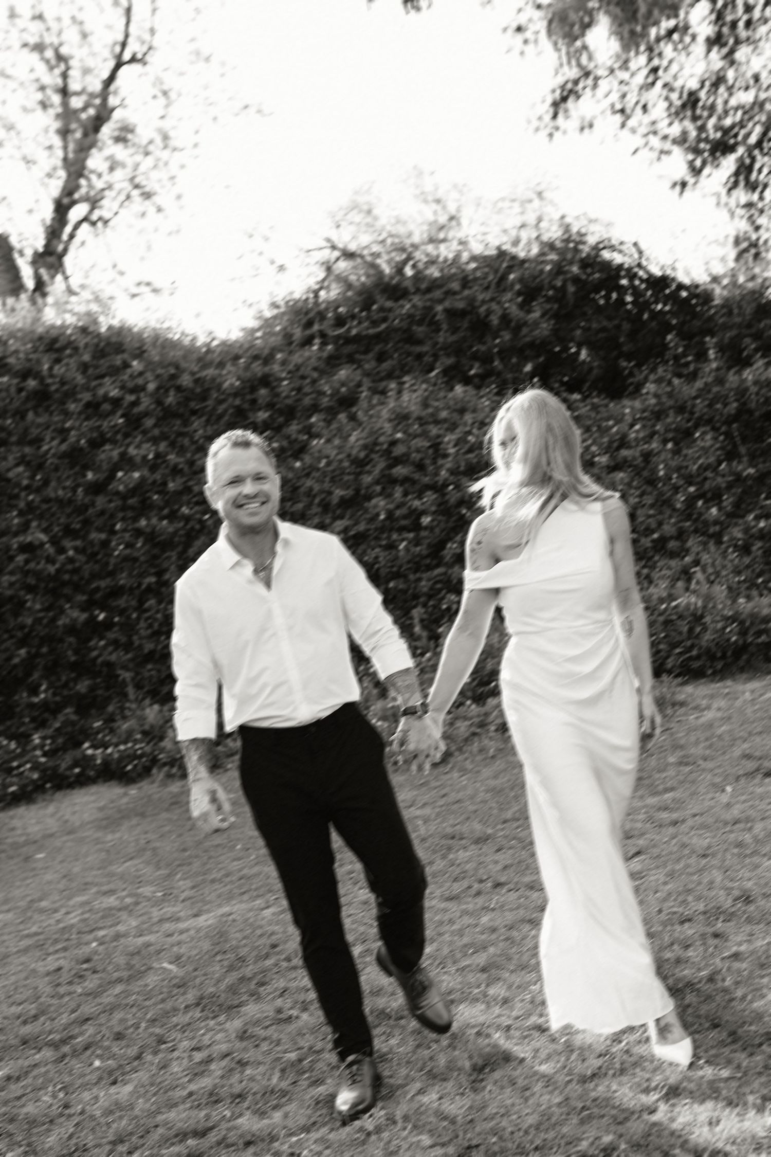 Black and white photo of a smiling couple holding hands, walking on grass with lush hedges in the background.