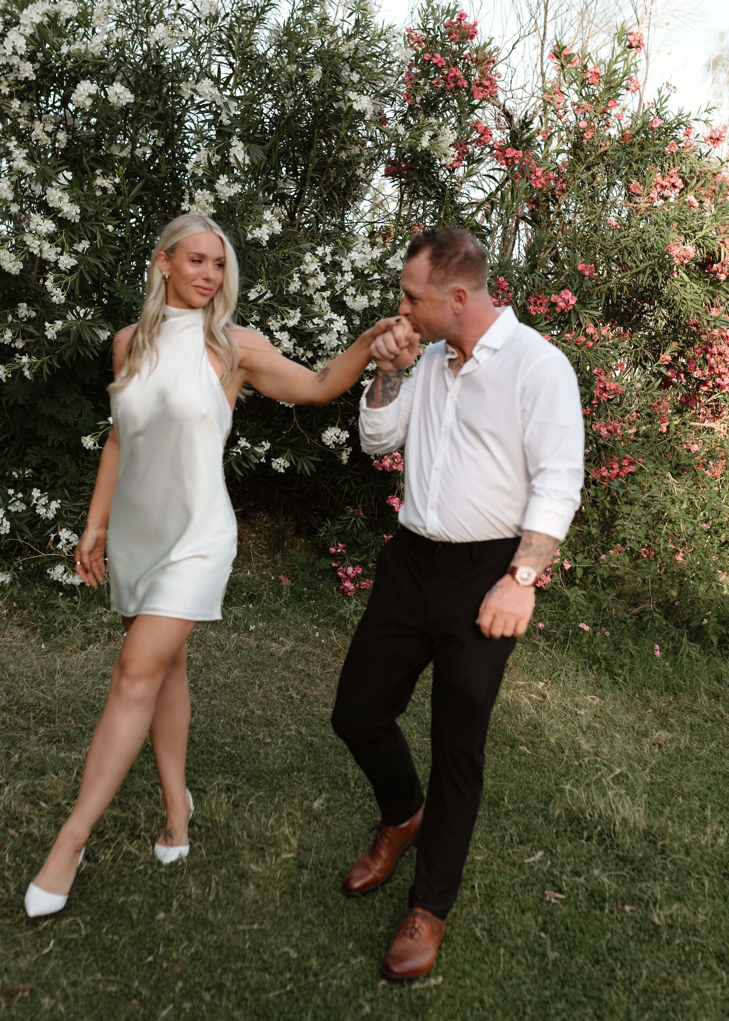 A man in a white shirt kisses a woman's hand as she smiles in a white dress, posing in a garden.