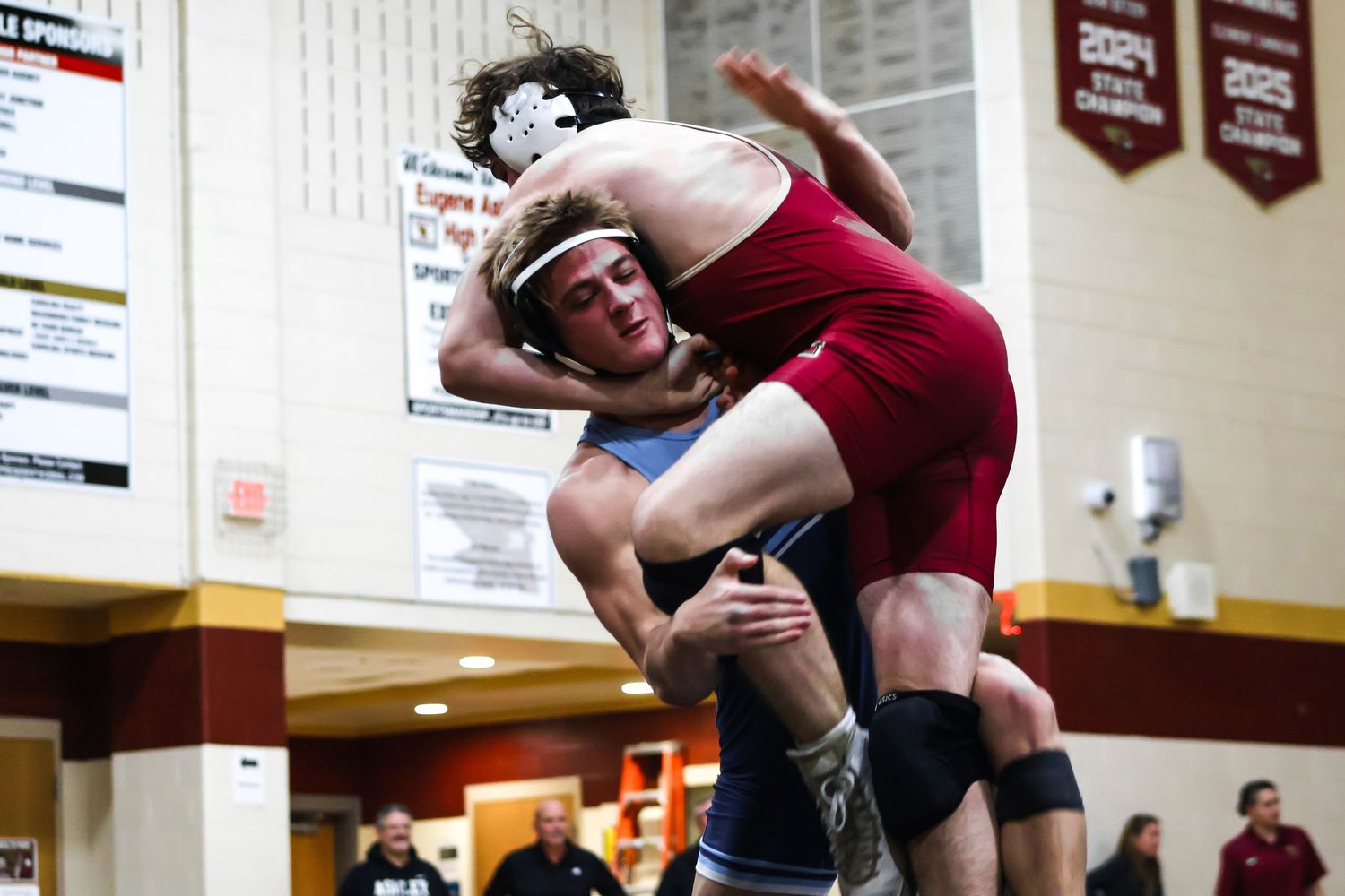 High school wrestlers compete in a wrestling match on a gymnasium mat as spectators watch from the sidelines.
