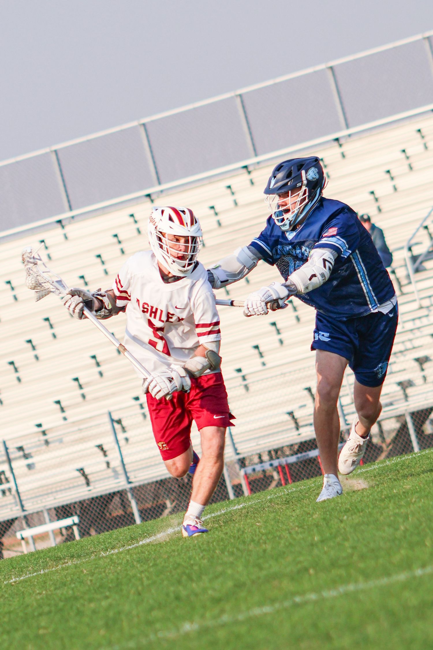 Two lacrosse players dive for the ball during an intense match on a grass field with stadium seating in background.