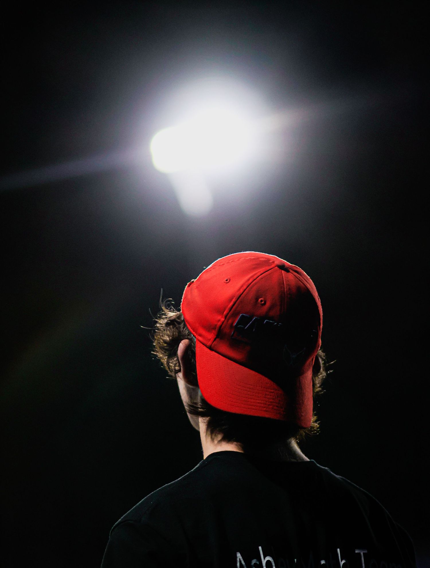 Person in red cap and black shirt viewed from behind, illuminated by bright spotlight against dark background.