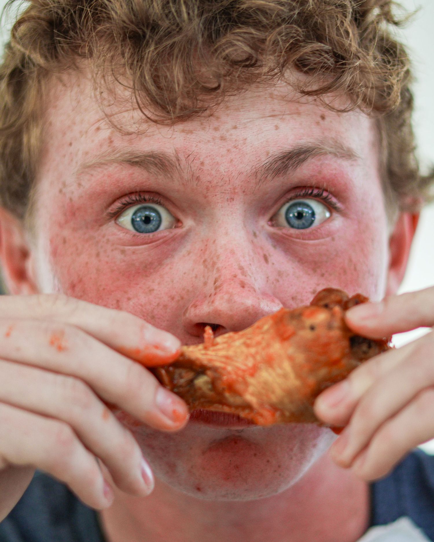 Freckled blue-eyed boy eating a saucy chicken wing, looking wide-eyed at camera.