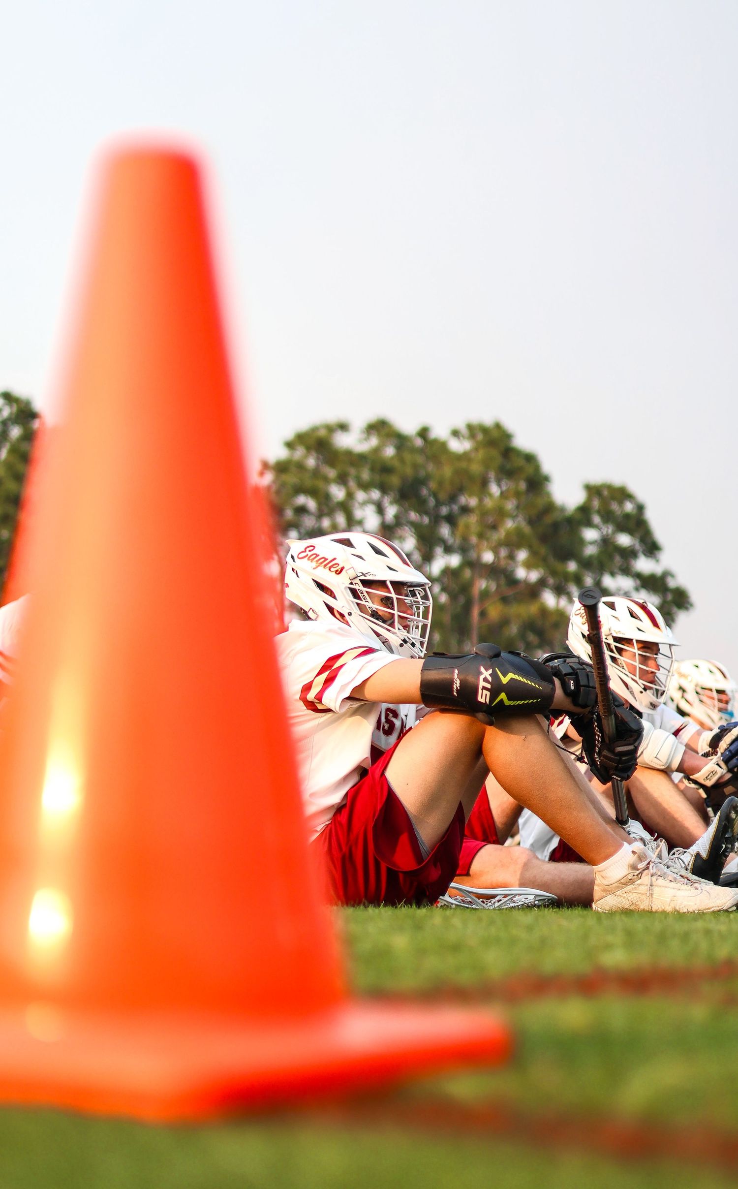 Orange sports cones in foreground with lacrosse players kneeling on grass field during practice session.