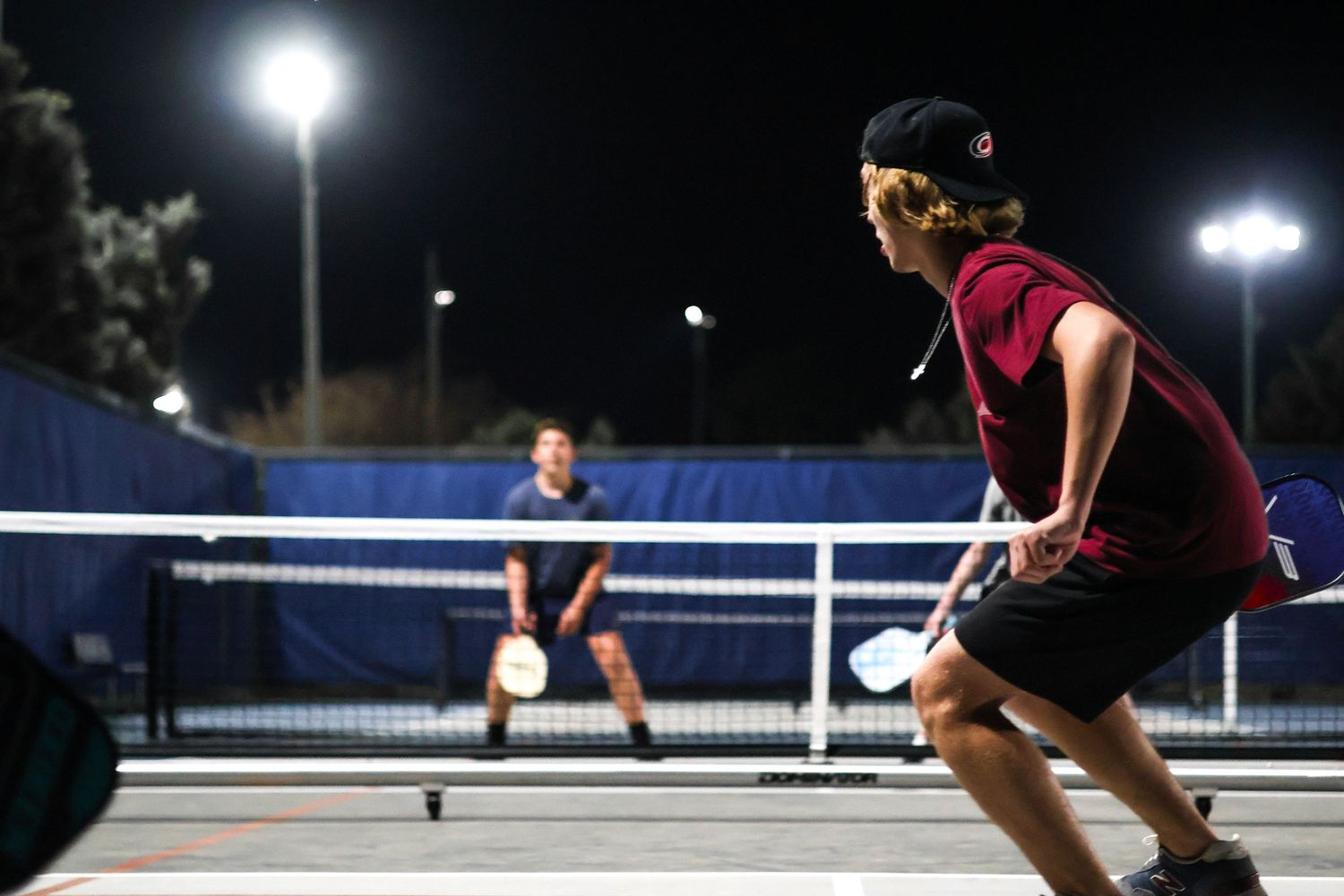 Two players compete in a nighttime pickleball match under bright court lights, with a net visible between them.