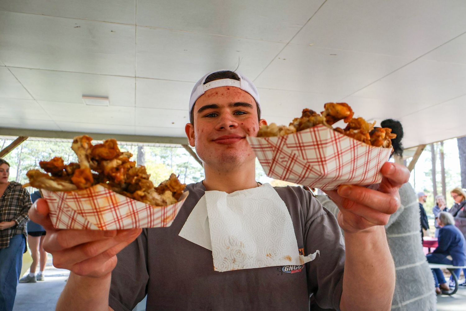 Young man in apron proudly holds two red checkered baskets of crispy fried food at a food festival.