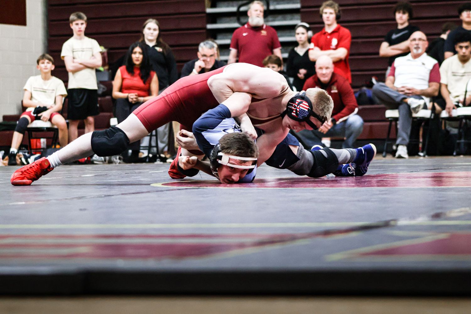 Two wrestlers compete on a mat during a match while spectators watch from the sidelines.