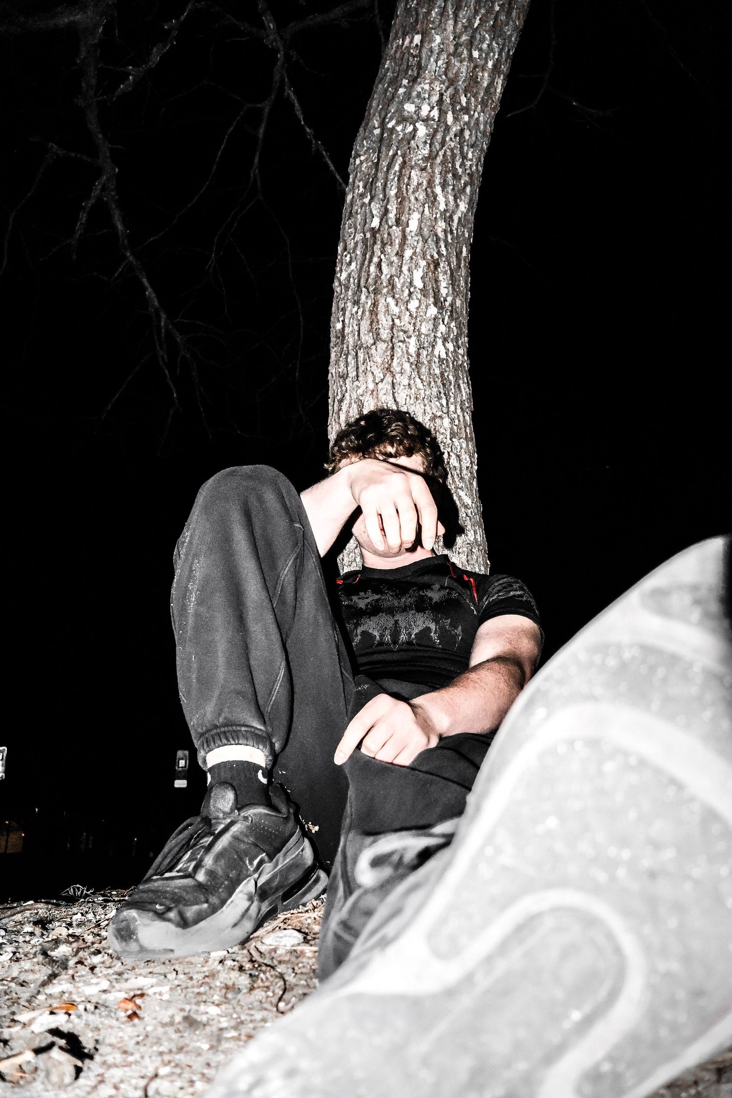 Young man sitting alone against a tree at night, head in hands, in a dark outdoor setting.