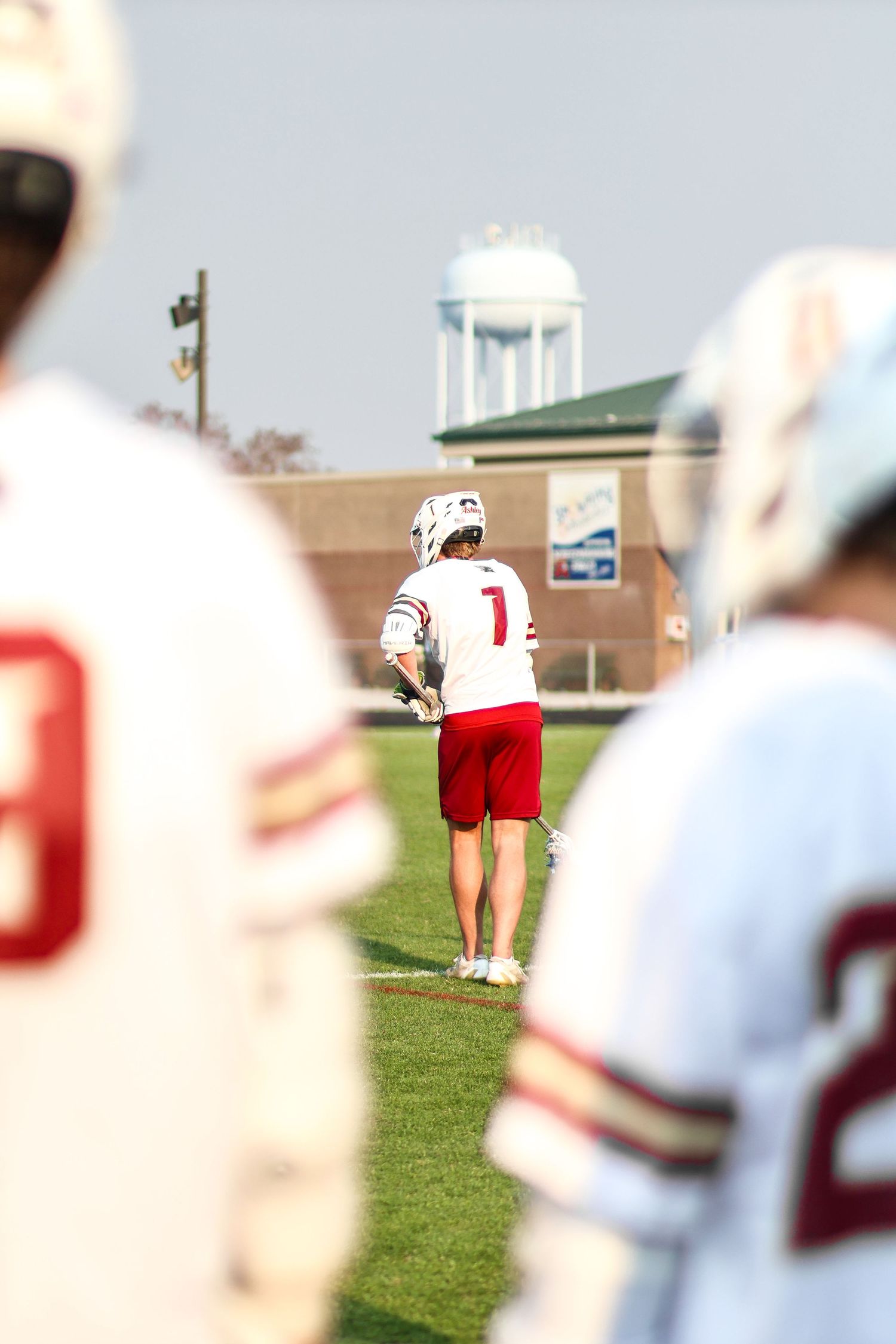 Football players in red and white uniforms stand on a sunny practice field with goal posts visible in the distance.
