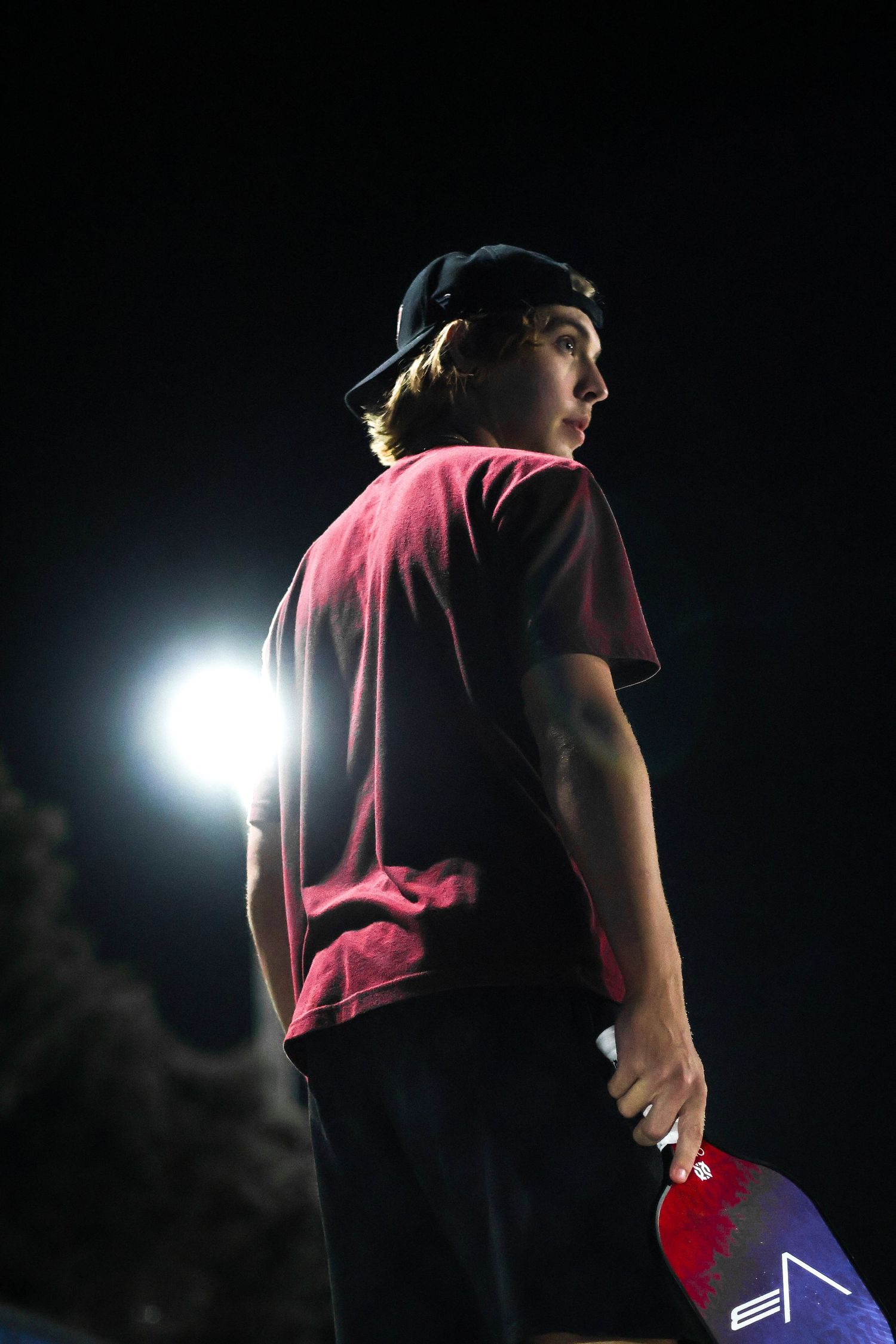 Young skateboarder in red shirt and backwards cap holding skateboard at night with bright light behind him.