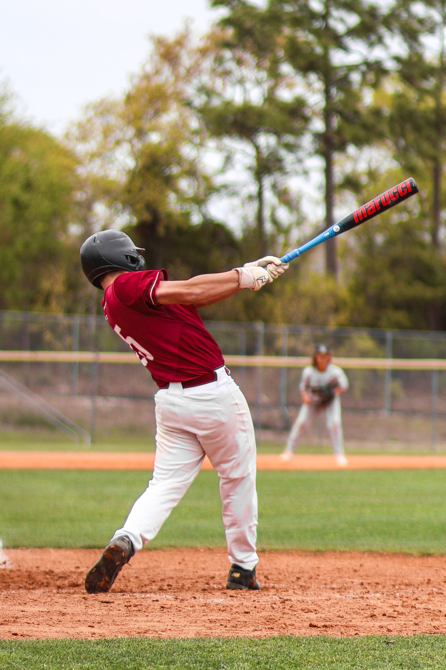 Baseball batter in red jersey swings at pitch during outdoor game, with fielder visible in background.