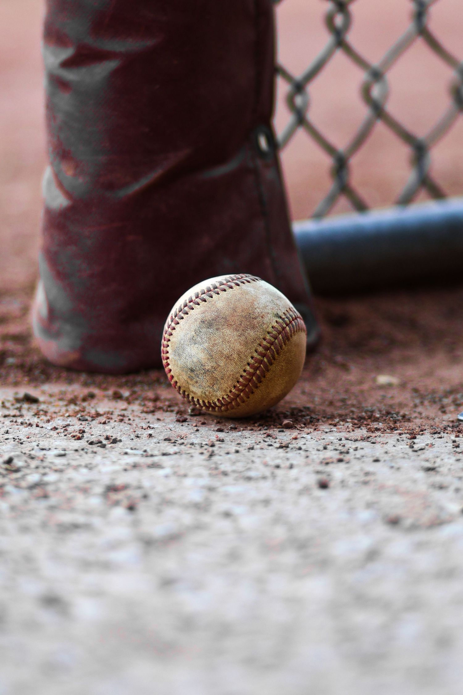 A worn baseball lies on dirt infield with chain link fence and cleats in the background.