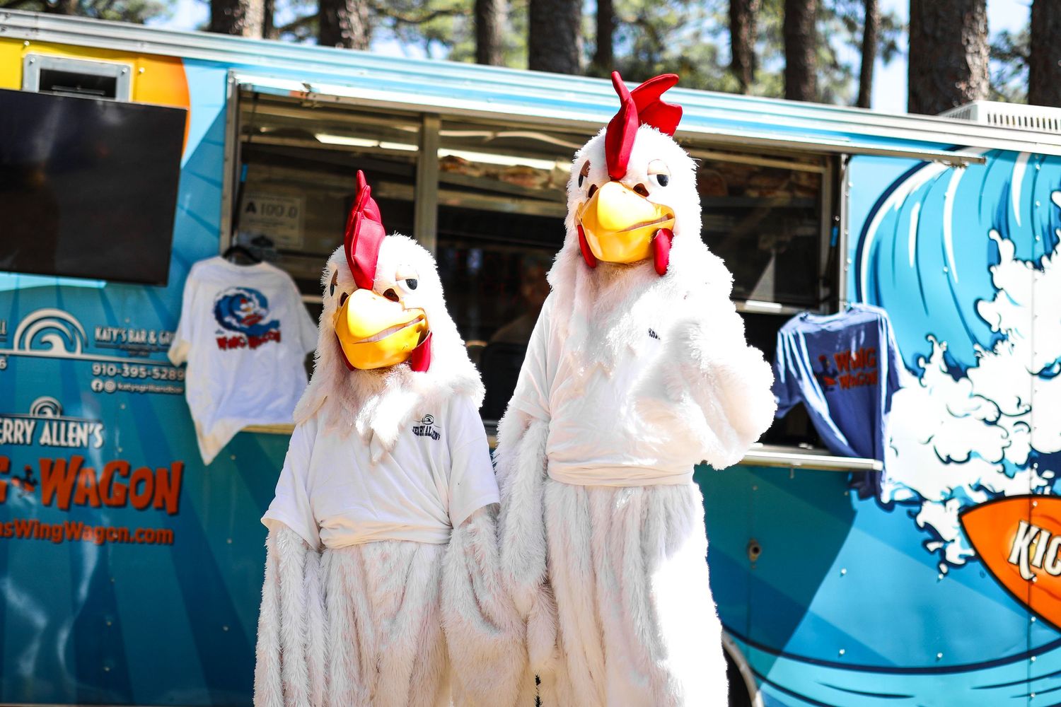 Two people in white rooster costumes pose in front of a colorful food truck at an outdoor event.