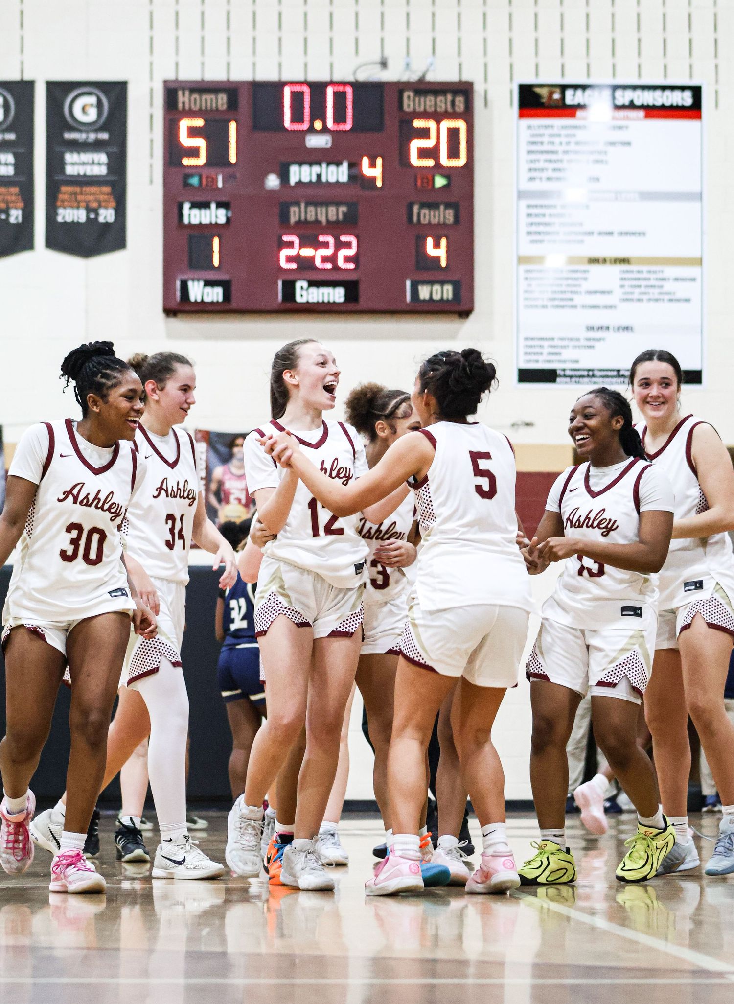 Basketball players in white jerseys line up on the sideline during a game with scoreboard visible in background.