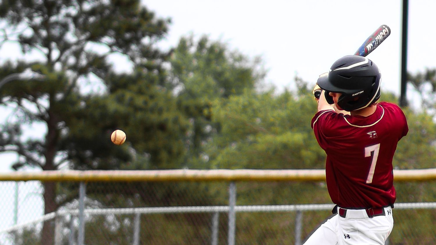 Baseball batter in maroon number 7 jersey swings at incoming pitch during a daytime baseball game.