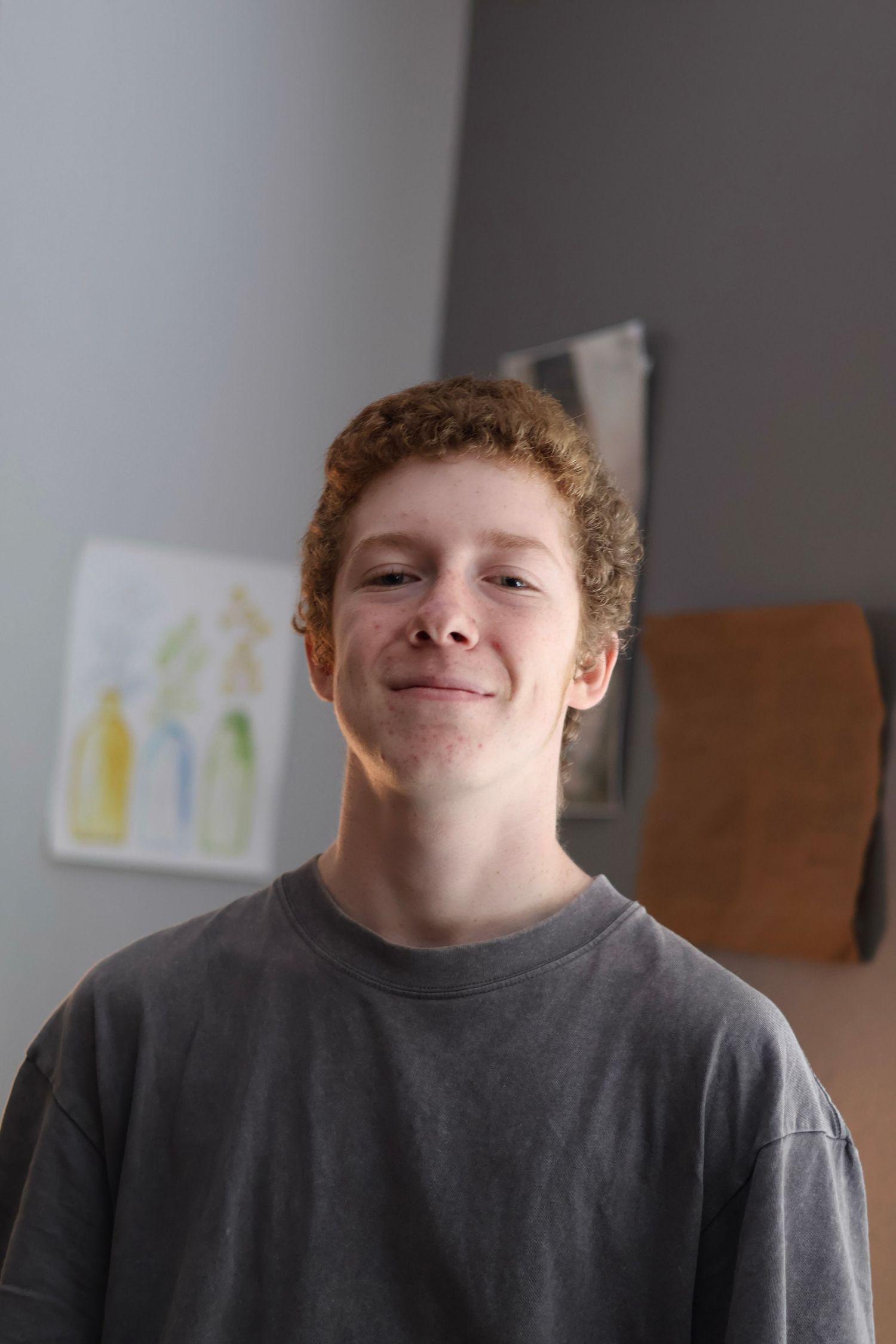 Smiling teenage boy with curly red hair wearing a gray t-shirt standing indoors near colorful artwork.