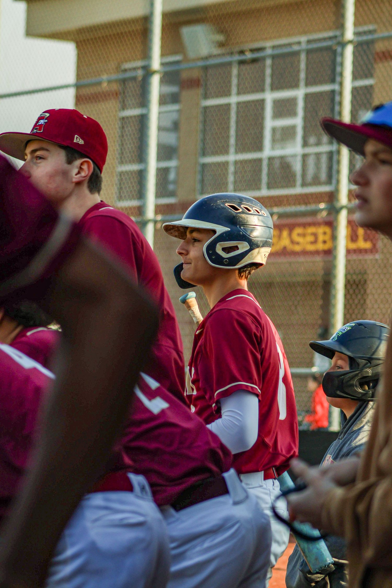 Baseball players in maroon uniforms gather in the dugout during a game with brick building visible in background.