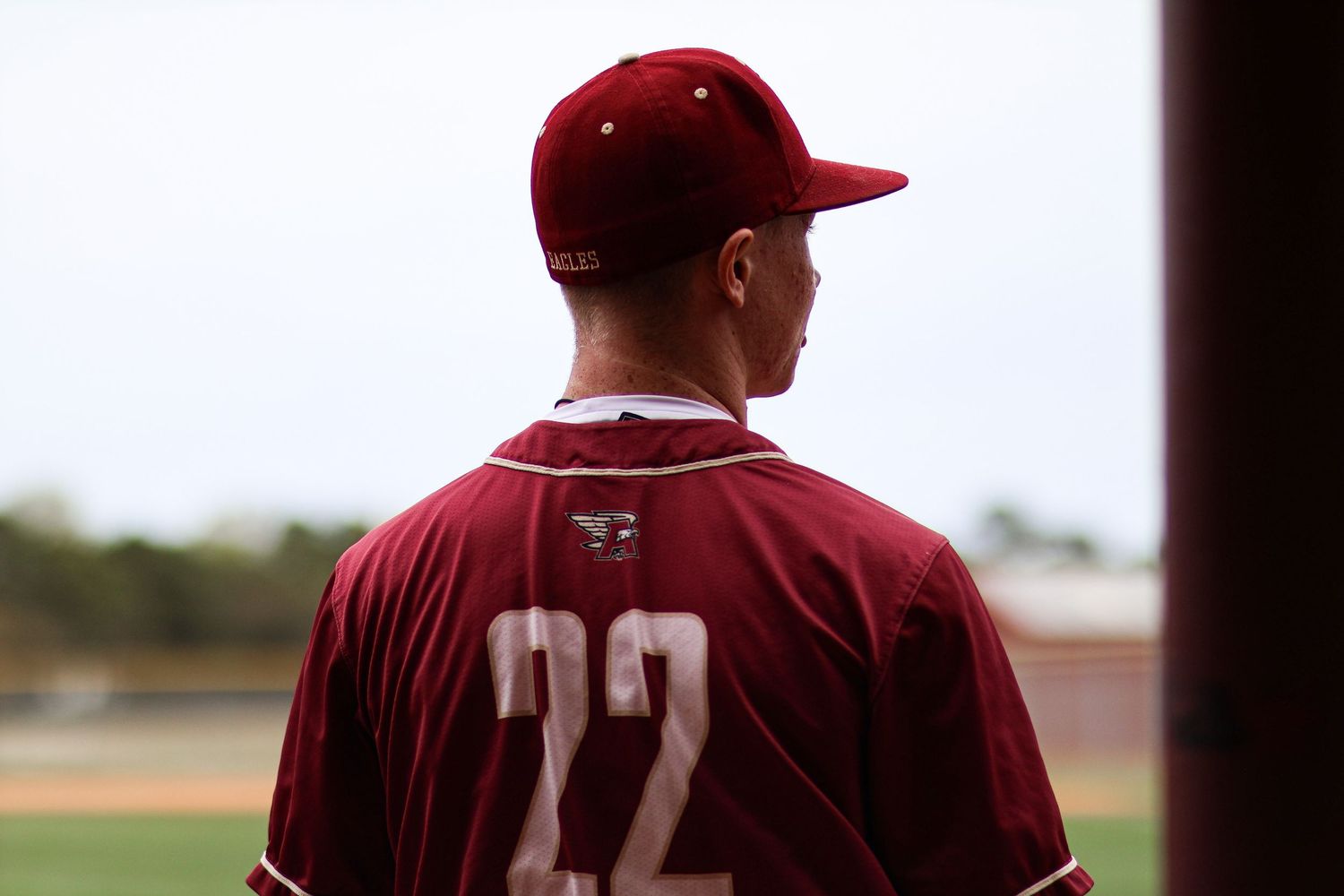 Baseball player wearing maroon number 22 jersey and cap stands with back to camera on a baseball field.