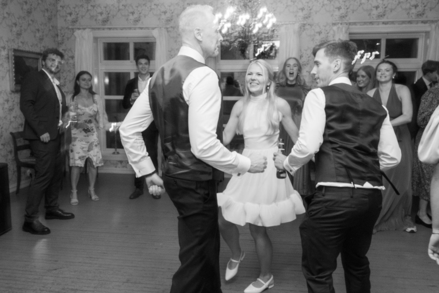 Bride in short white dress dancing joyfully with guests on wedding reception dance floor.