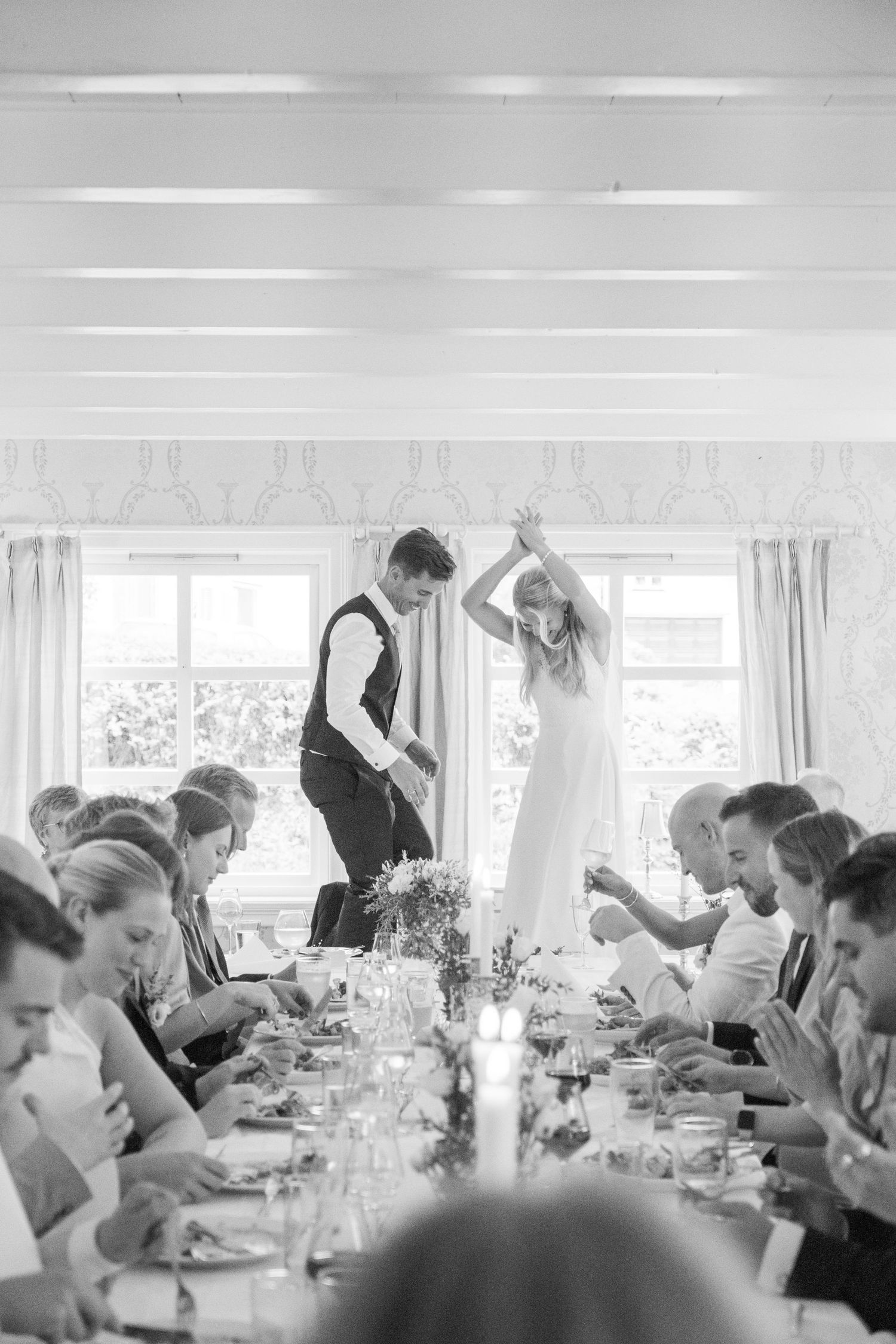 Black and white reception photo of bride and groom dancing joyfully at a long candlelit dinner table.