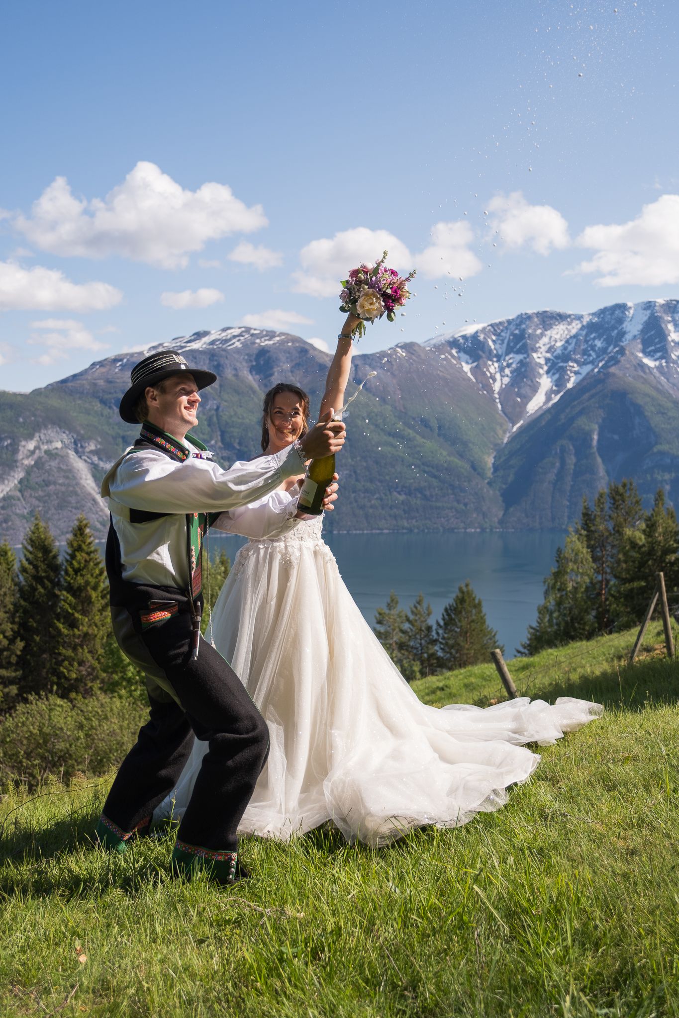 Joyful bride and groom in Norwegian bunad popping champagne bottle on a green hillside with fjord and mountains behind.