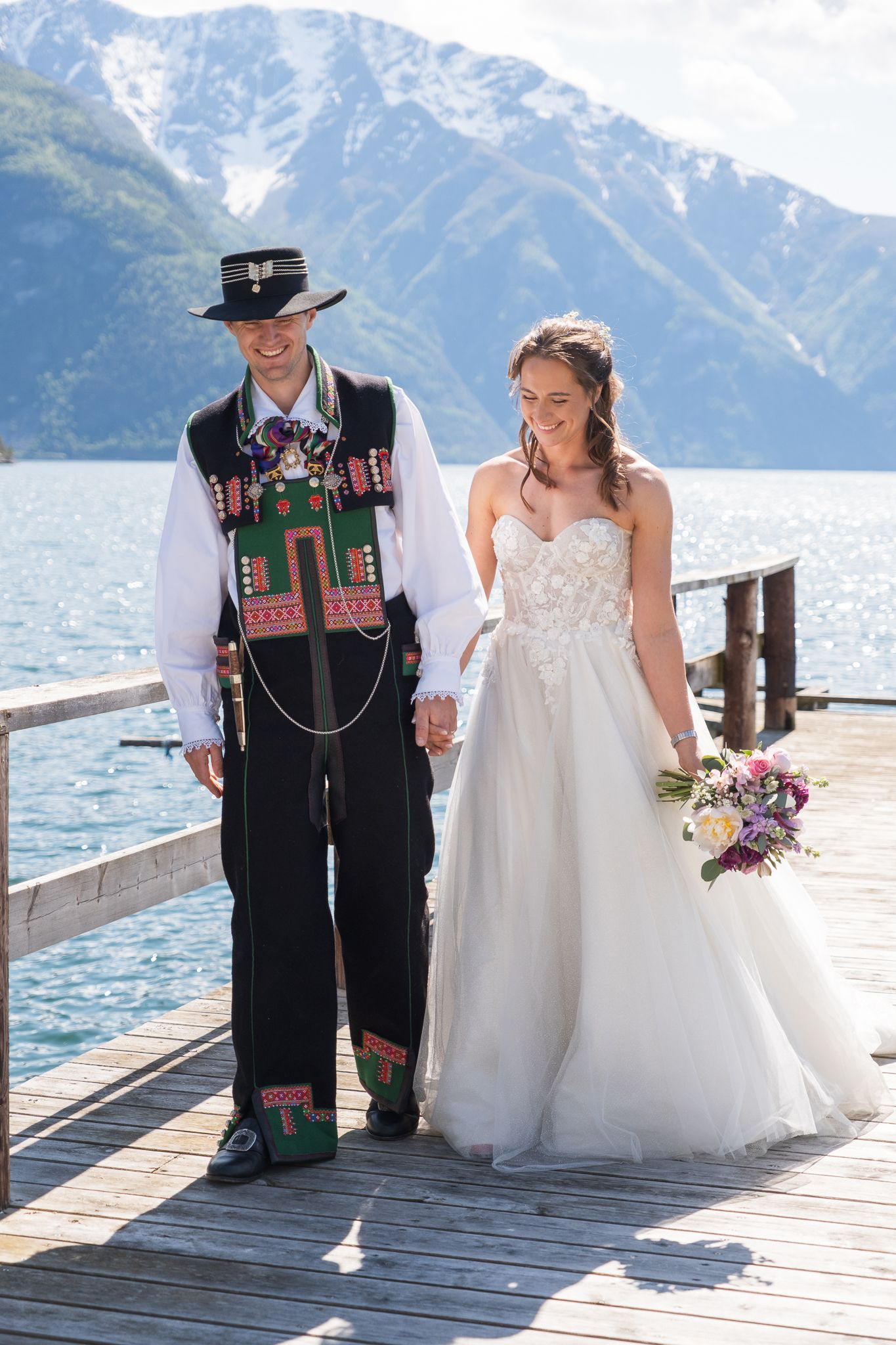 Groom in traditional folk costume walks with bride in white gown on a lakeside dock with mountains.