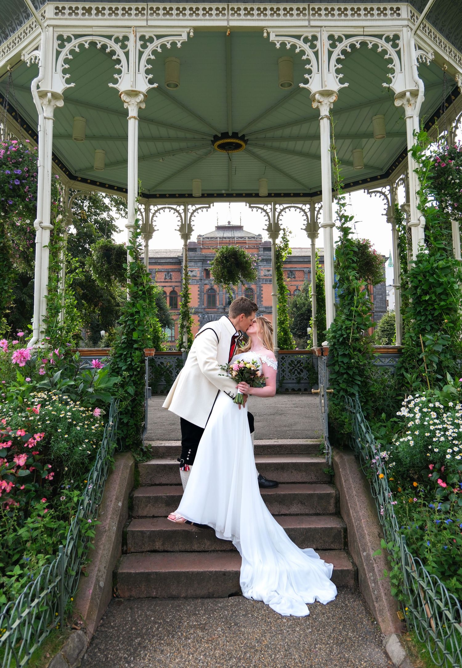 Bride and groom sharing a kiss on steps of ornate Victorian bandstand surrounded by colorful flowers.