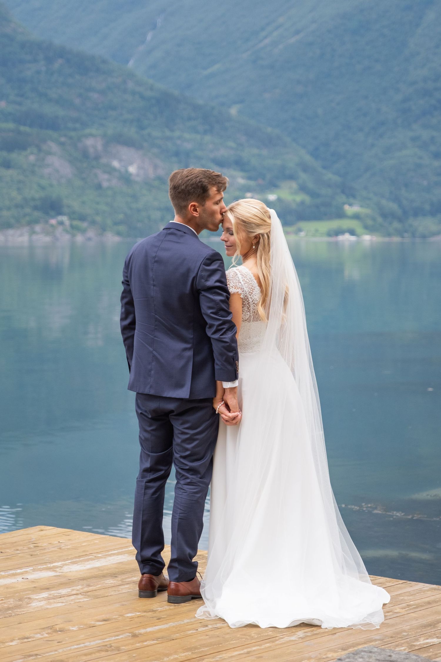 Bride and groom holding hands on wooden dock overlooking serene fjord and mountains.