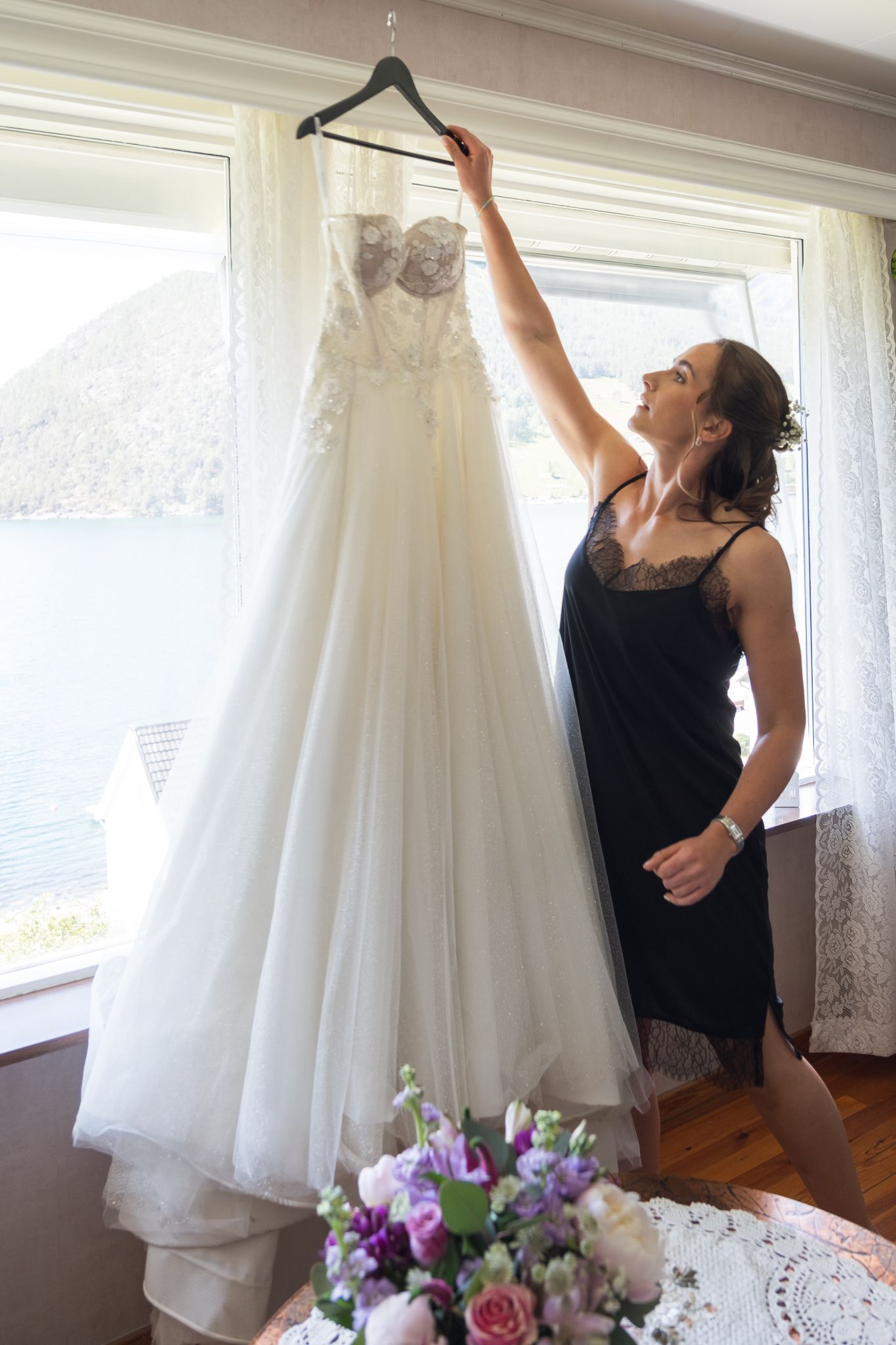 Woman in black dress reaches up to admire strapless lace wedding gown hanging by bright window.