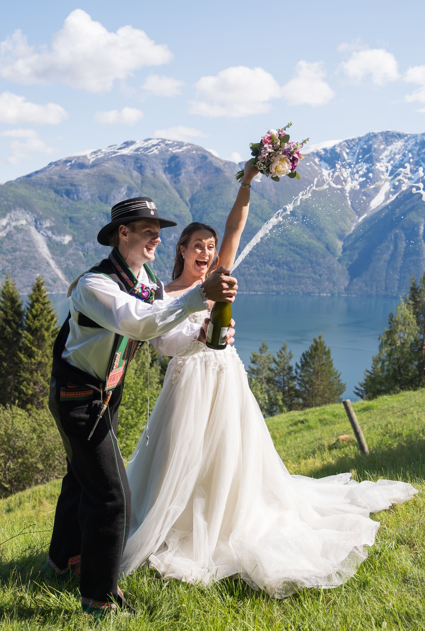 Joyful newlyweds popping champagne on a green hillside with a breathtaking fjord and mountain backdrop.