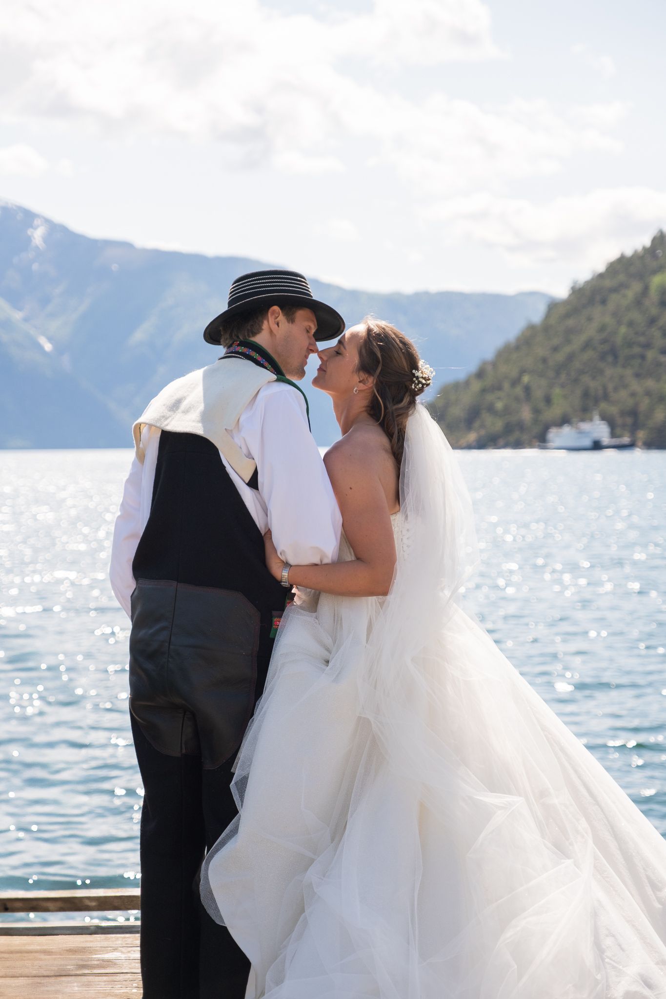 Bride and groom share a romantic kiss on a wooden pier with a sparkling mountain lake behind them.