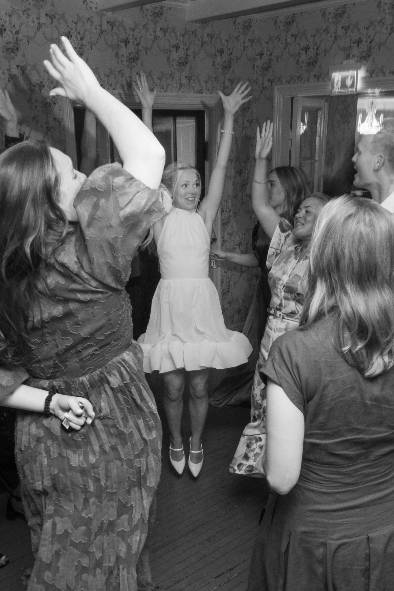 Black and white photo of bride in short dress jumping joyfully on the dance floor with wedding guests.